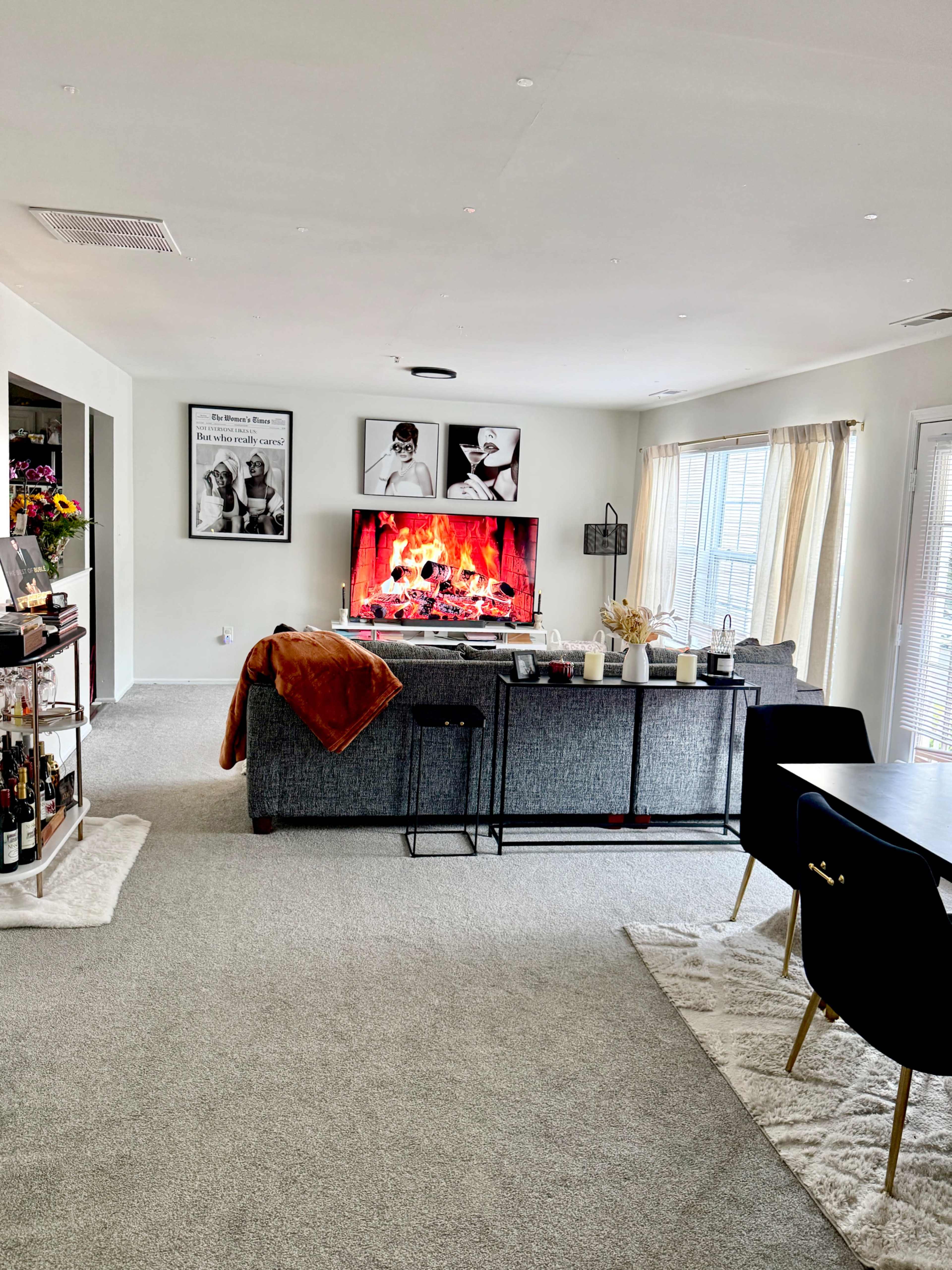 A cozy living room features a gray sofa facing a large TV displaying a fire, with a dining area visible on the right and decorative wall art on display.