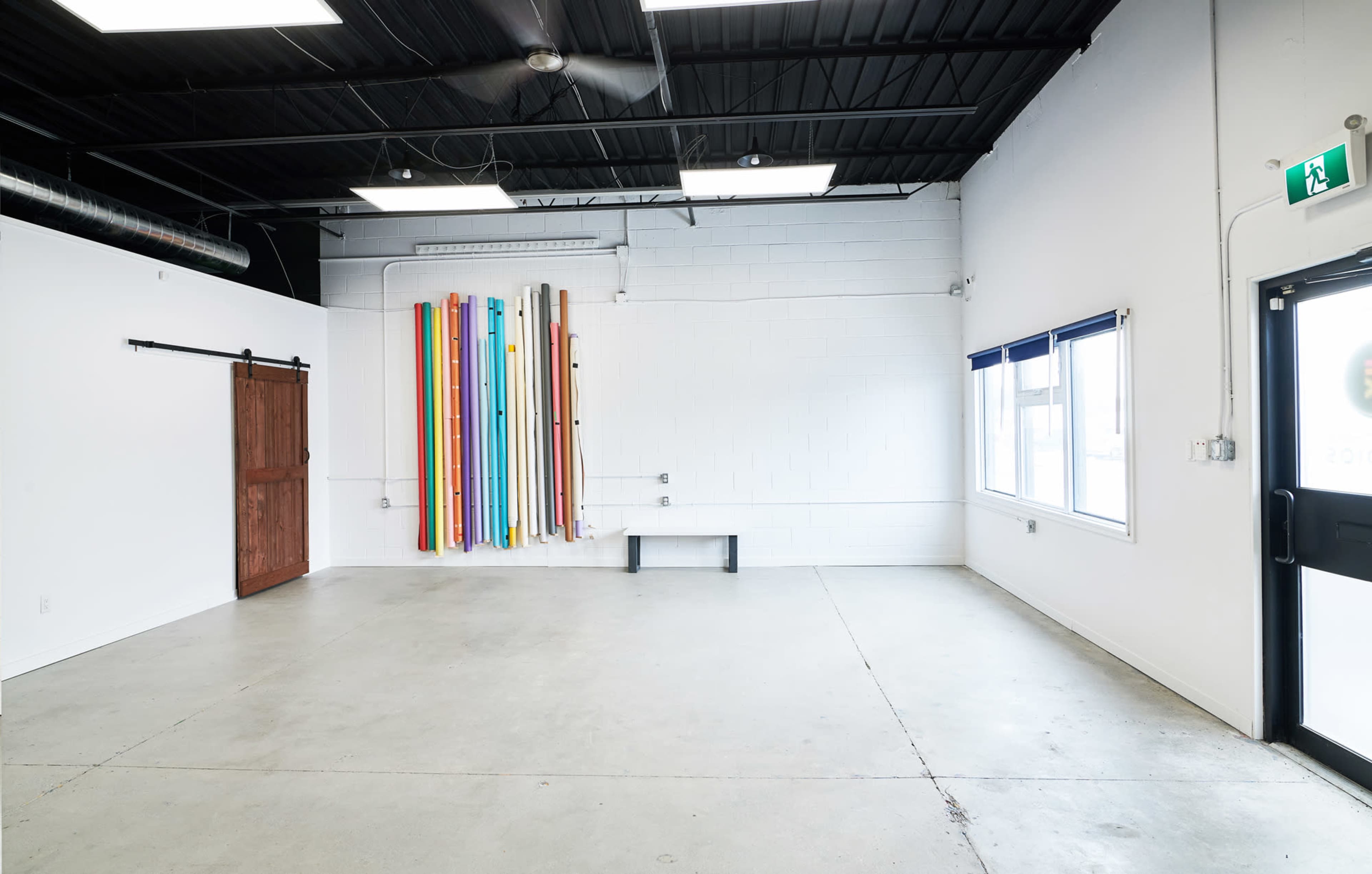 An empty room with a concrete floor, a wooden sliding door, colorful backdrops hanging on the wall, and large windows letting in natural light.