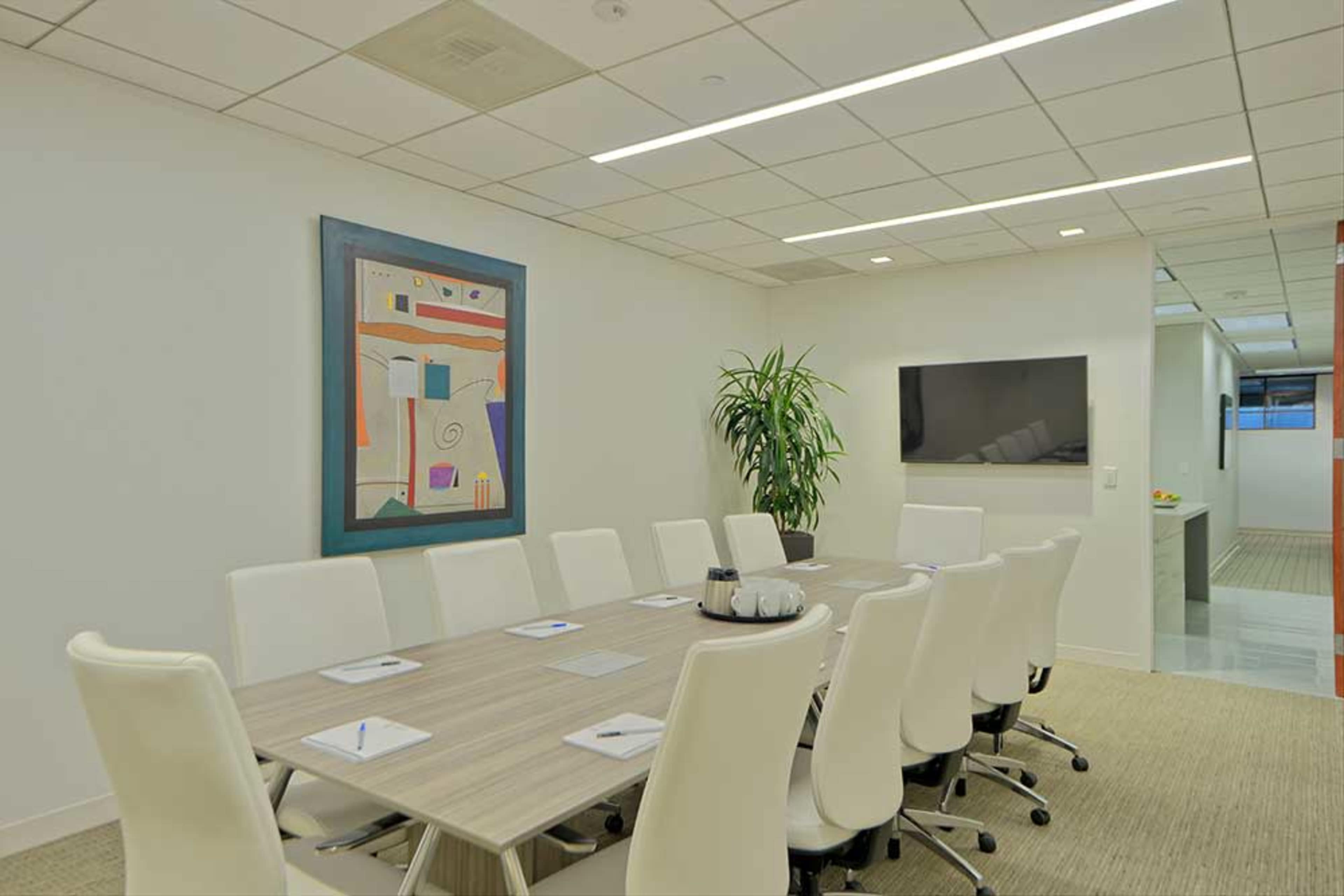 A modern conference room features a large oval table surrounded by white chairs, a wall-mounted TV, and a colorful abstract painting.