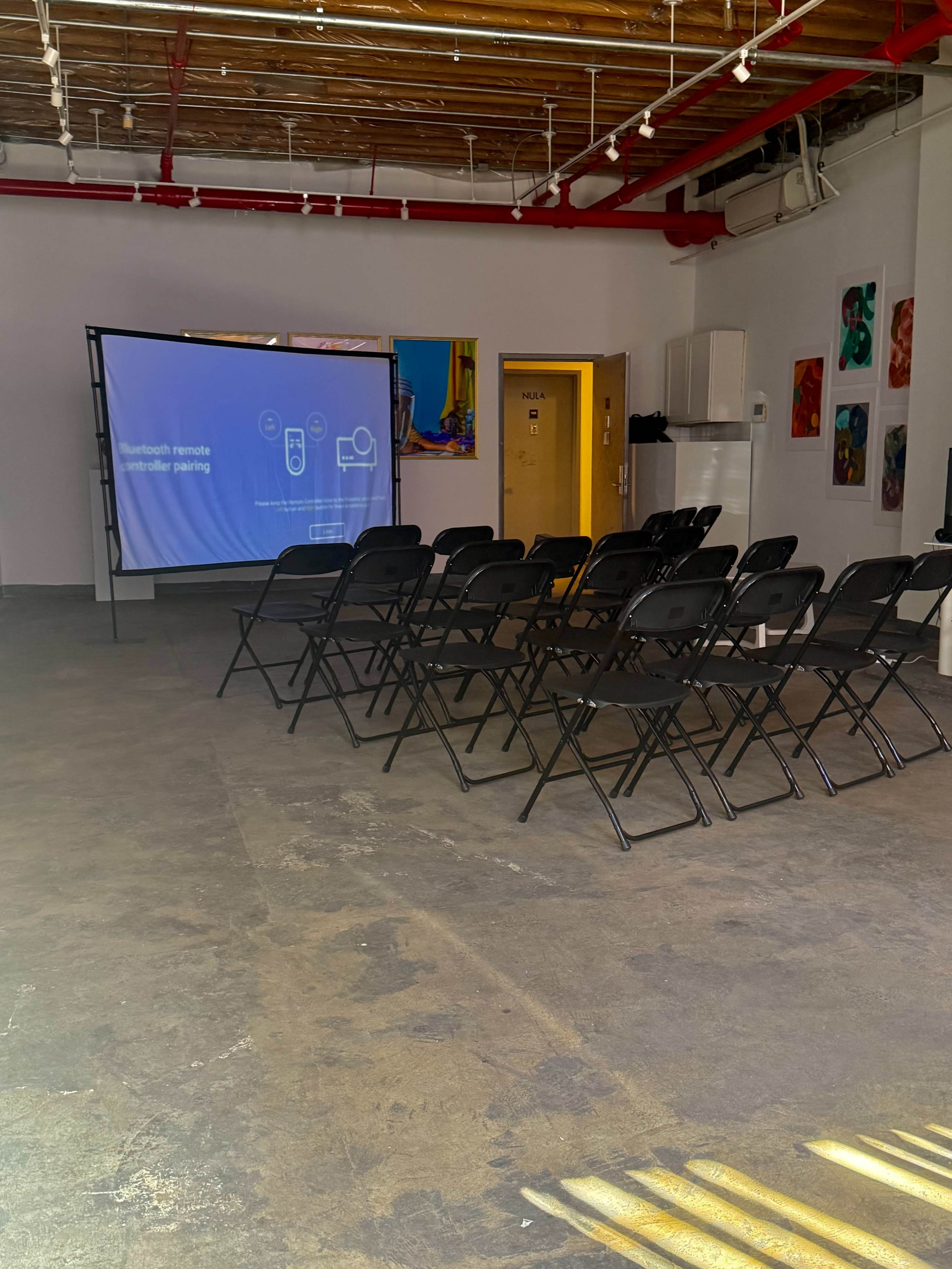 A row of black folding chairs faces a projector screen in a brightly lit room featuring colorful artworks on the walls.