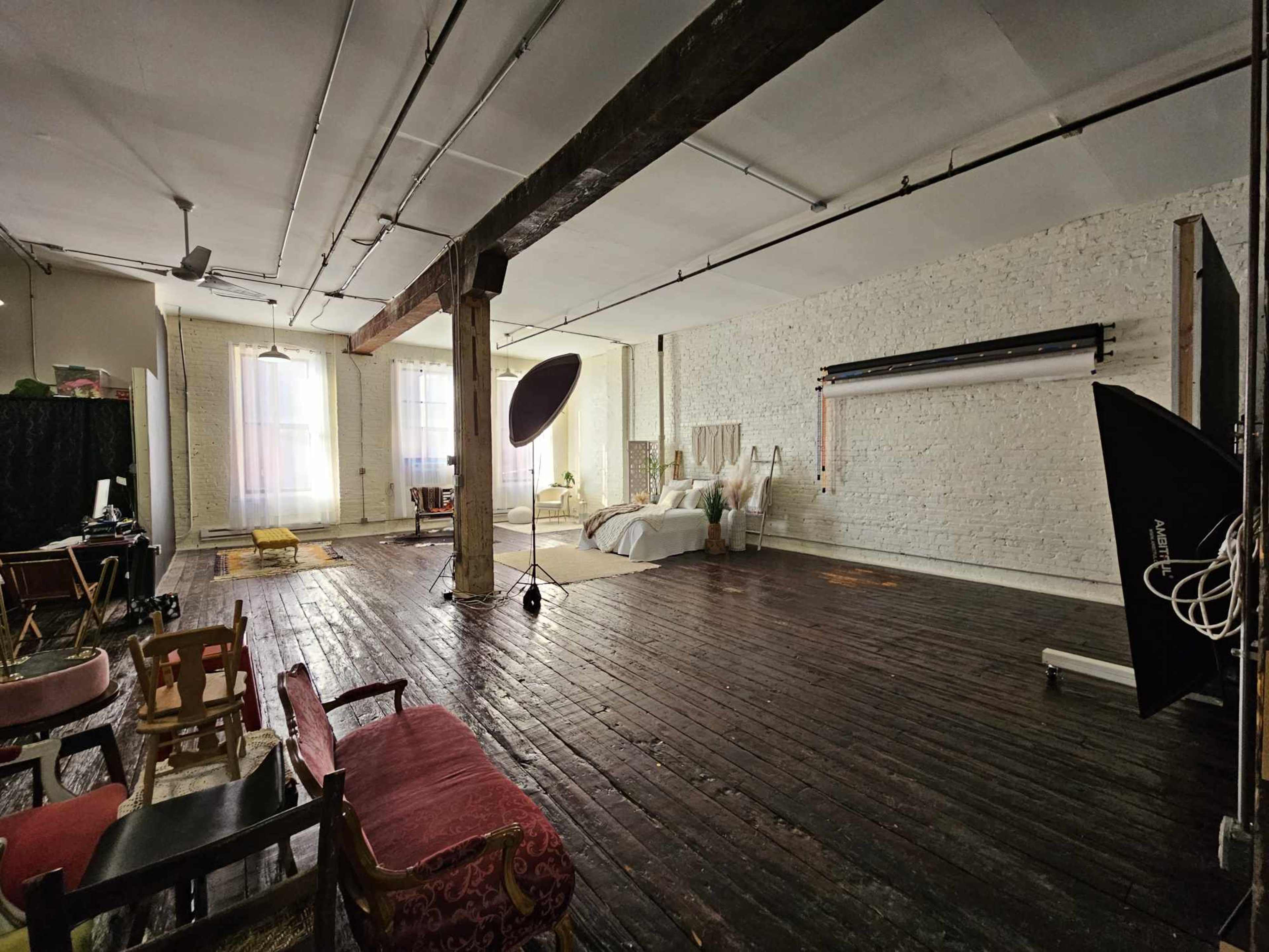 The image shows a spacious studio with wooden floors, white brick walls, large windows, and various furnishings, including a bed and photography equipment.