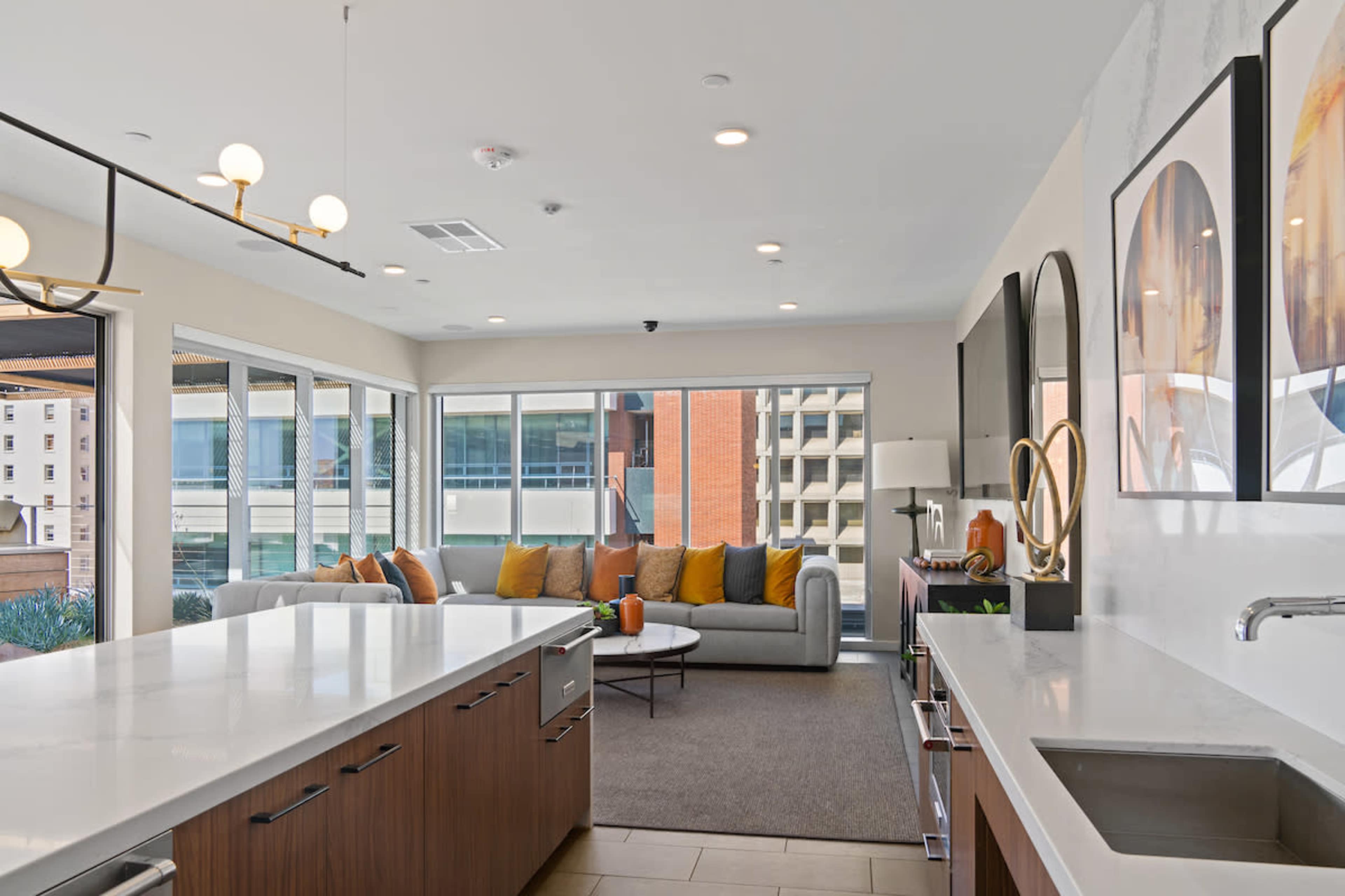 The image shows a modern kitchen with a large island, connected to a living area featuring a gray sofa and large windows allowing natural light.
