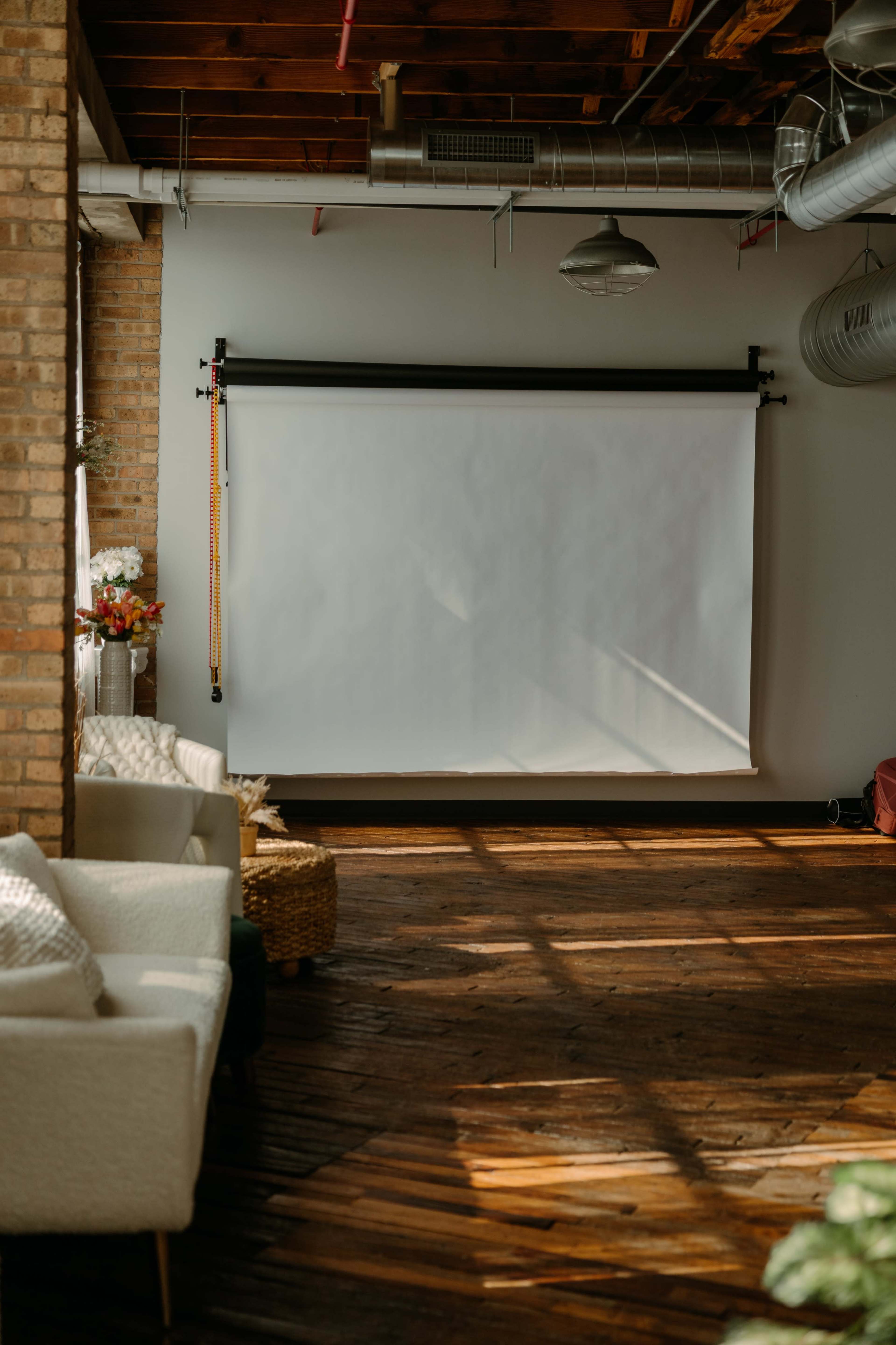 The image shows a spacious room with a blank projector screen mounted on a wall, hardwood flooring, and natural light casting shadows across the space.