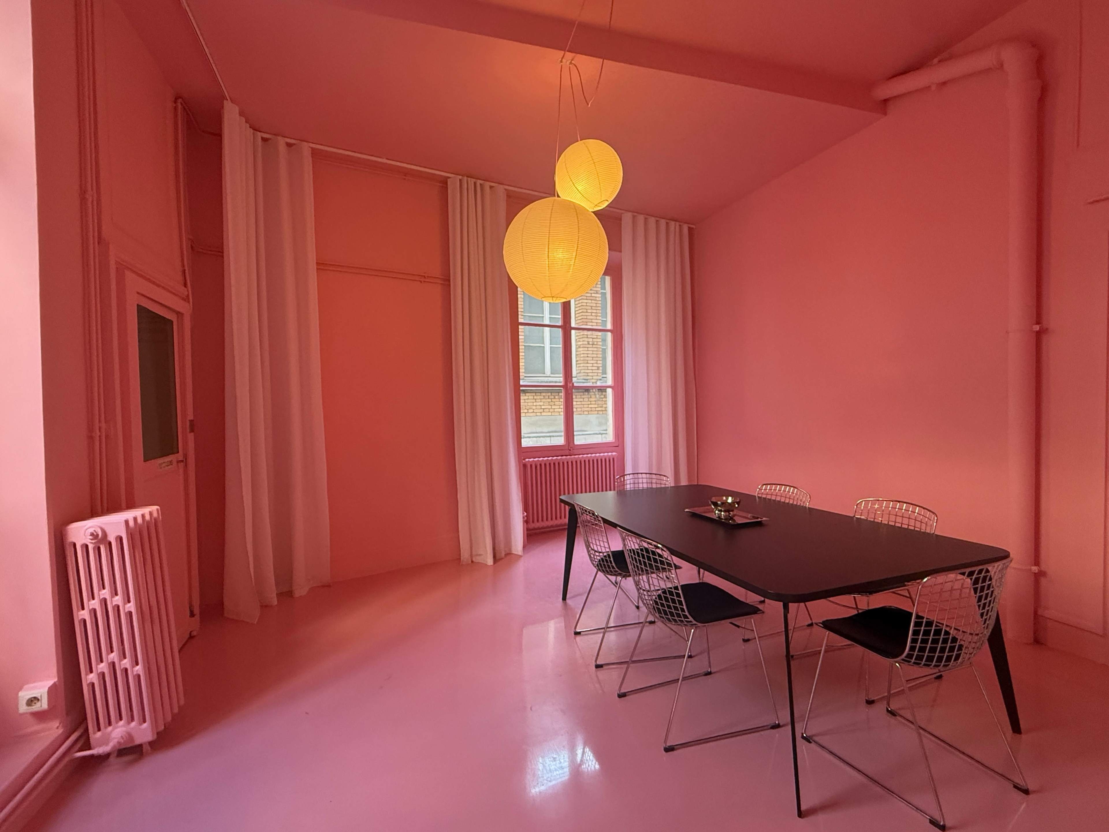 The image shows a pink room featuring a long black table surrounded by wire chairs, with two round pendant lamps hanging from the ceiling.