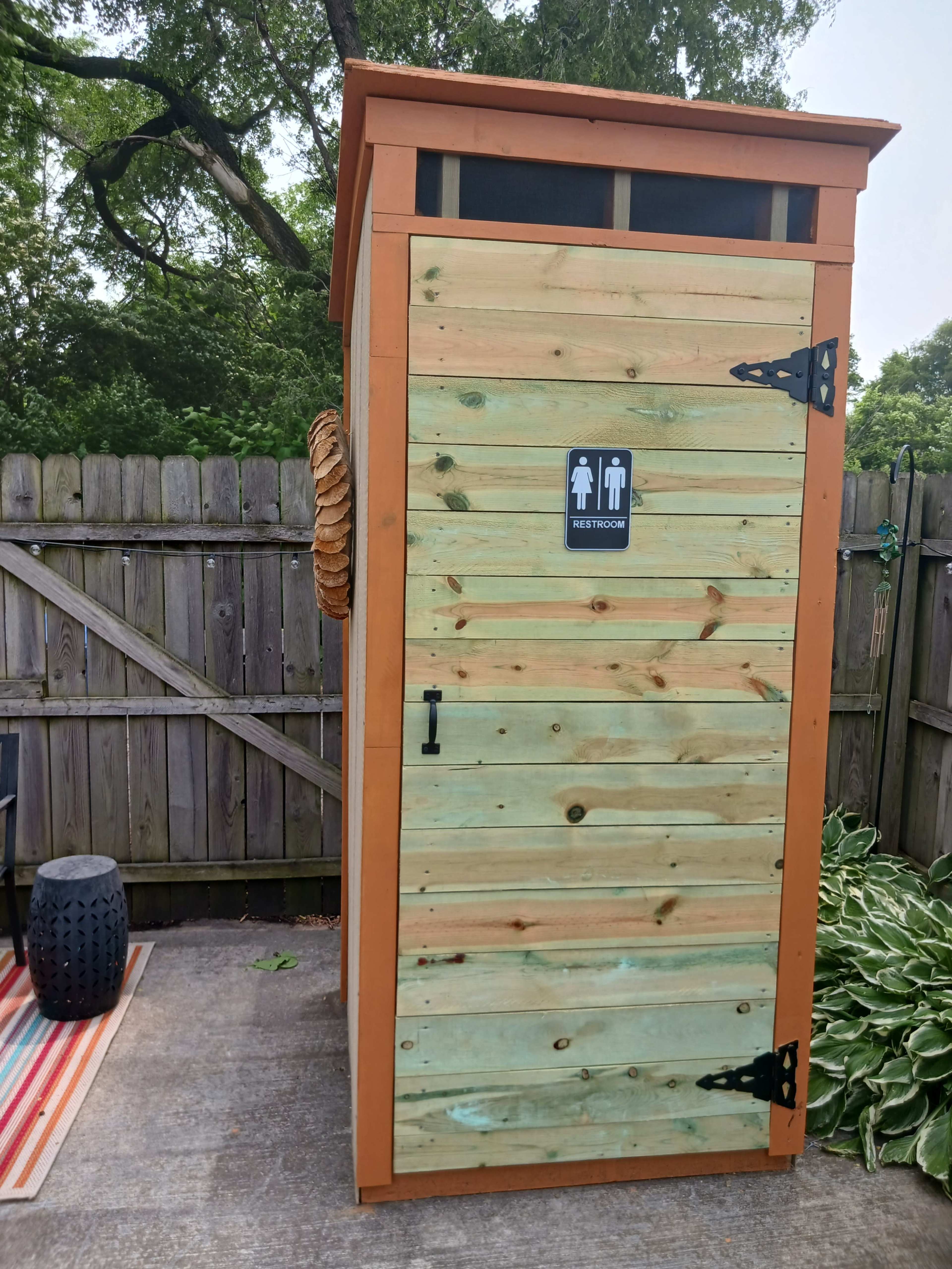 A wooden outdoor restroom with a paneled door, marked with a "Restroom" sign, set in a backyard area surrounded by fencing and landscaping.