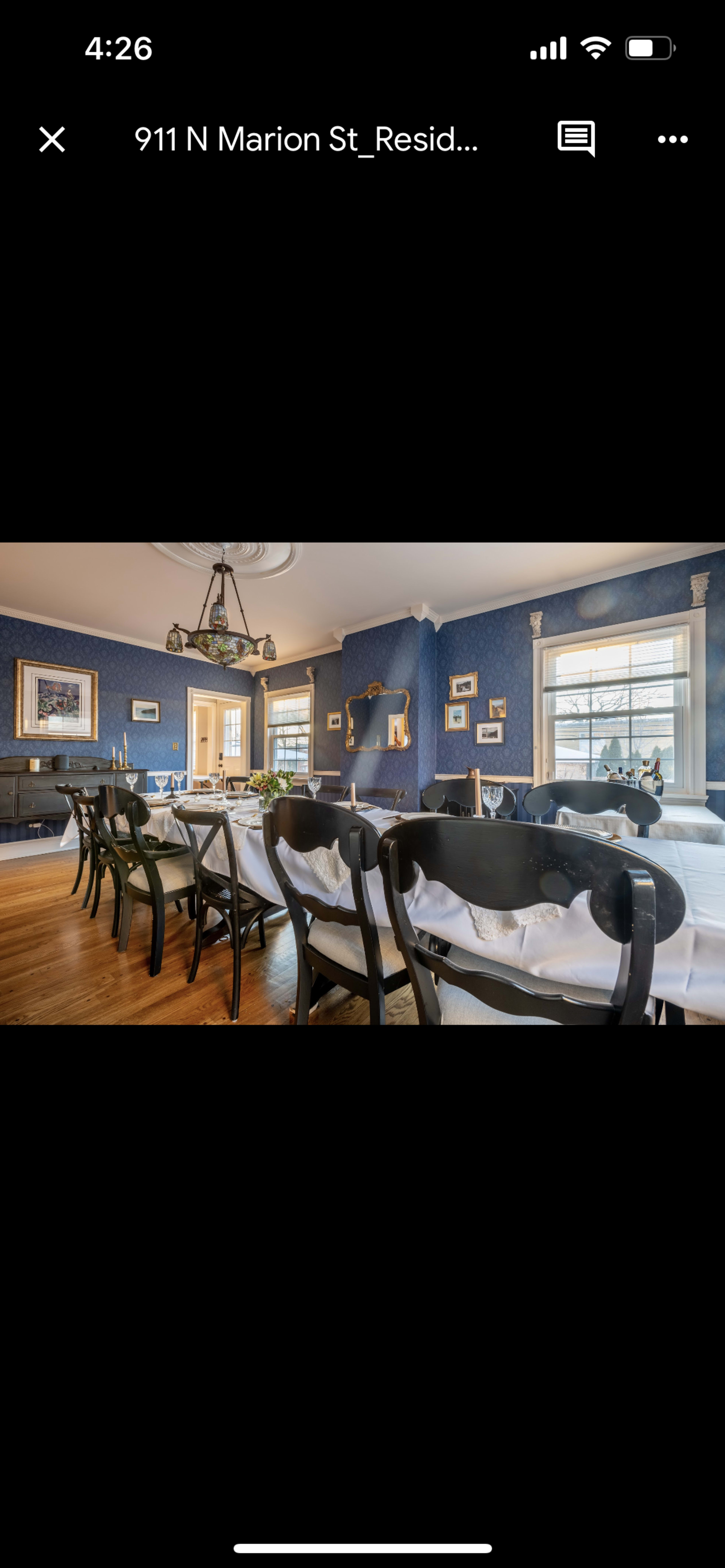 A dining room with a long table set with white tableware, surrounded by black chairs, and blue wallpaper adorned with framed artwork.