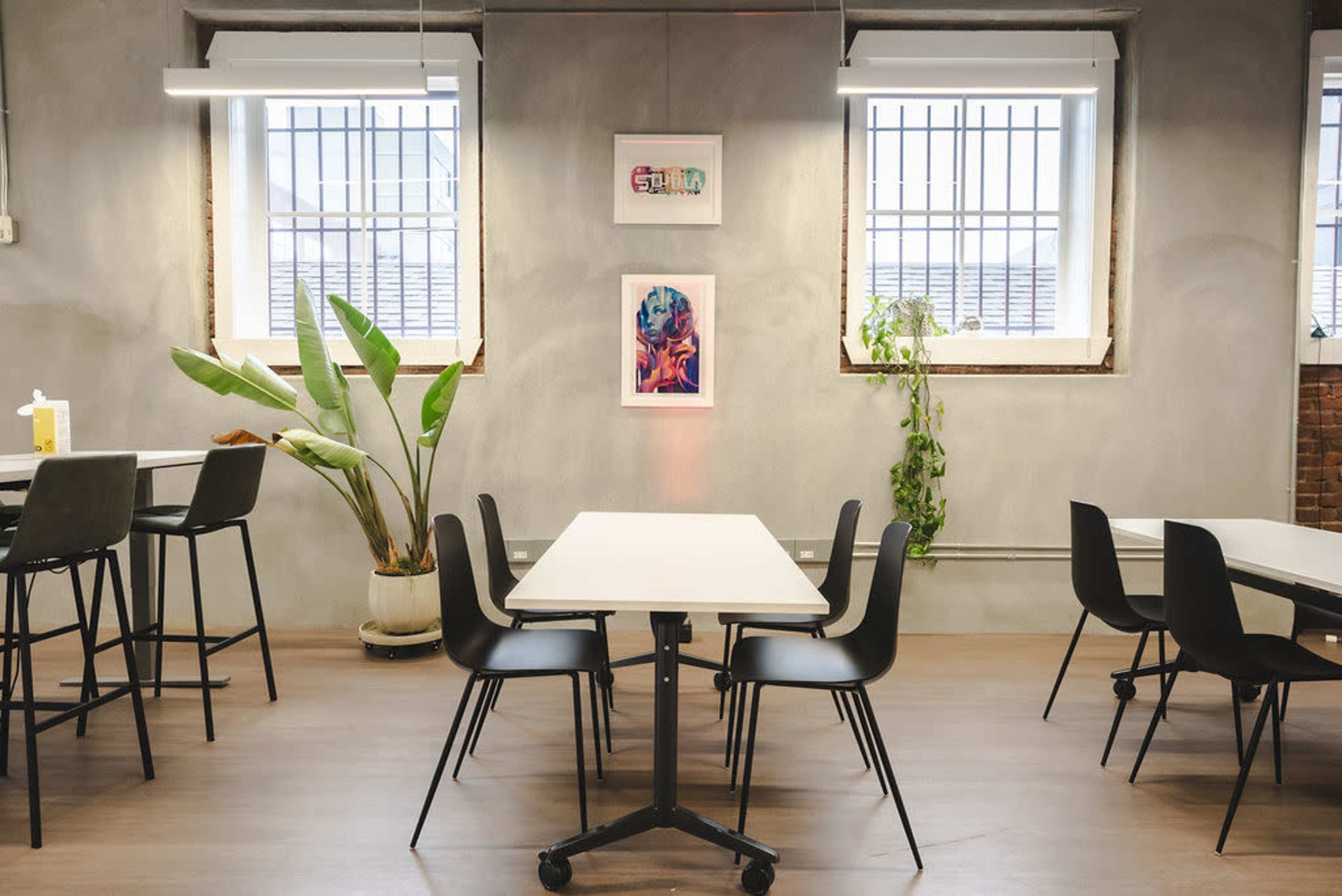 The image shows a modern, minimalistic workspace with several black chairs and tables arranged around a potted plant, featuring artwork on the walls and large windows.