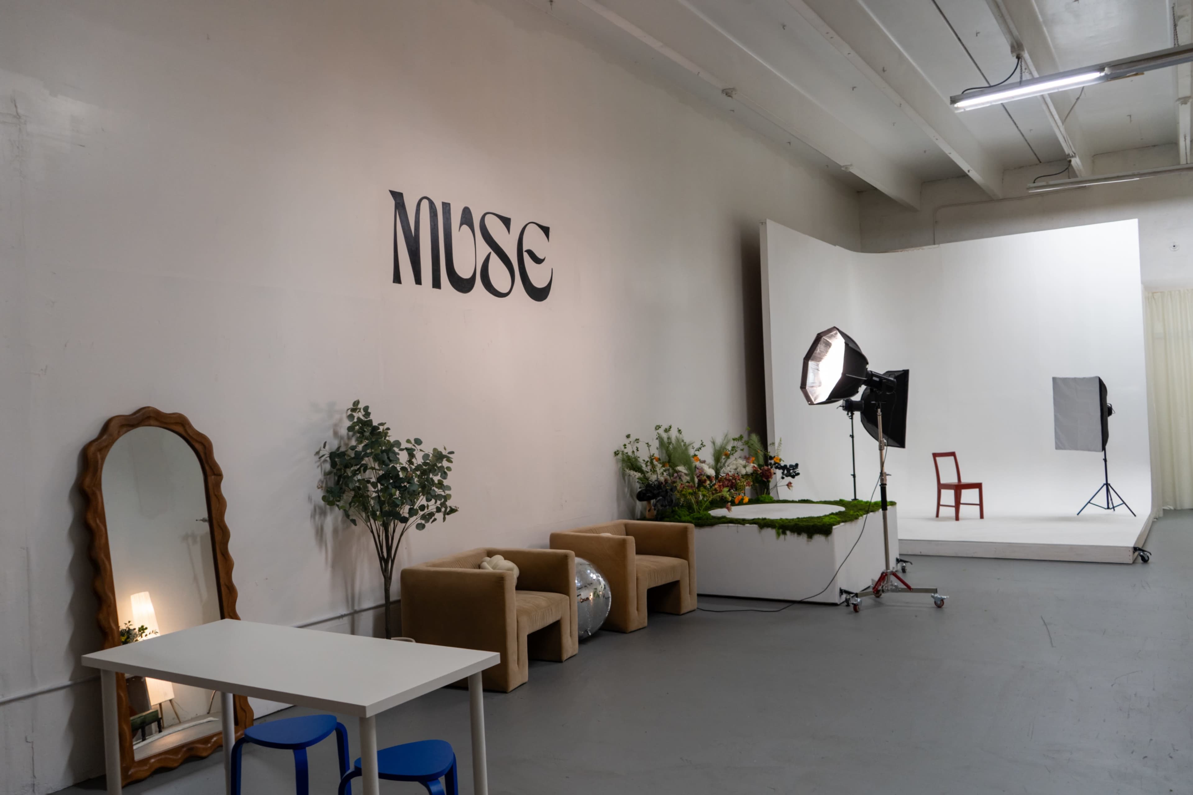The image shows a creatively designed studio space featuring a large "MUSE" sign, a mirror, a table, and photography equipment set against a minimalist backdrop.