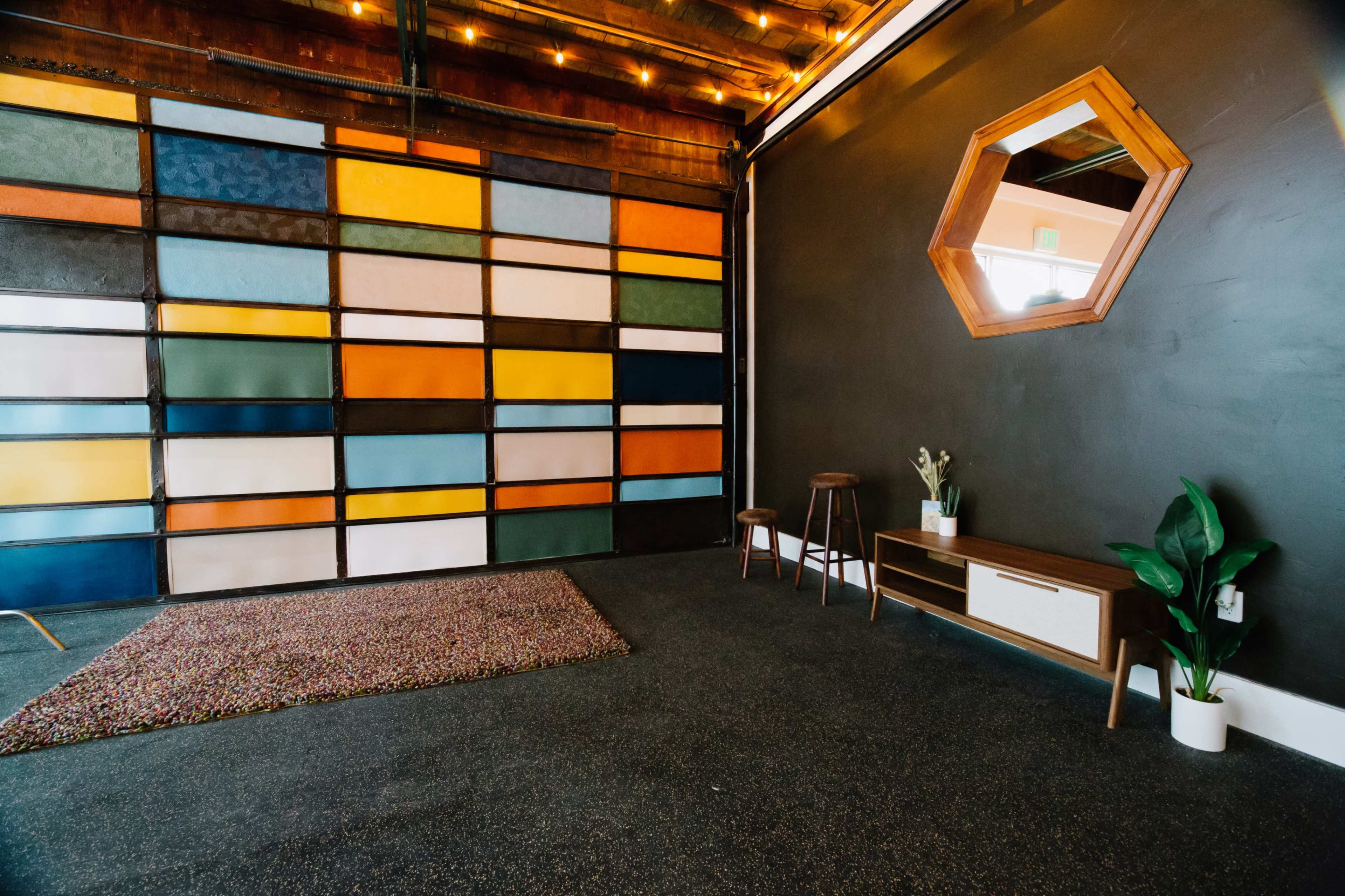 An interior space featuring a colorfully patterned wall, a dark floor, a mirror, a small wooden table, a plant, and a rug.