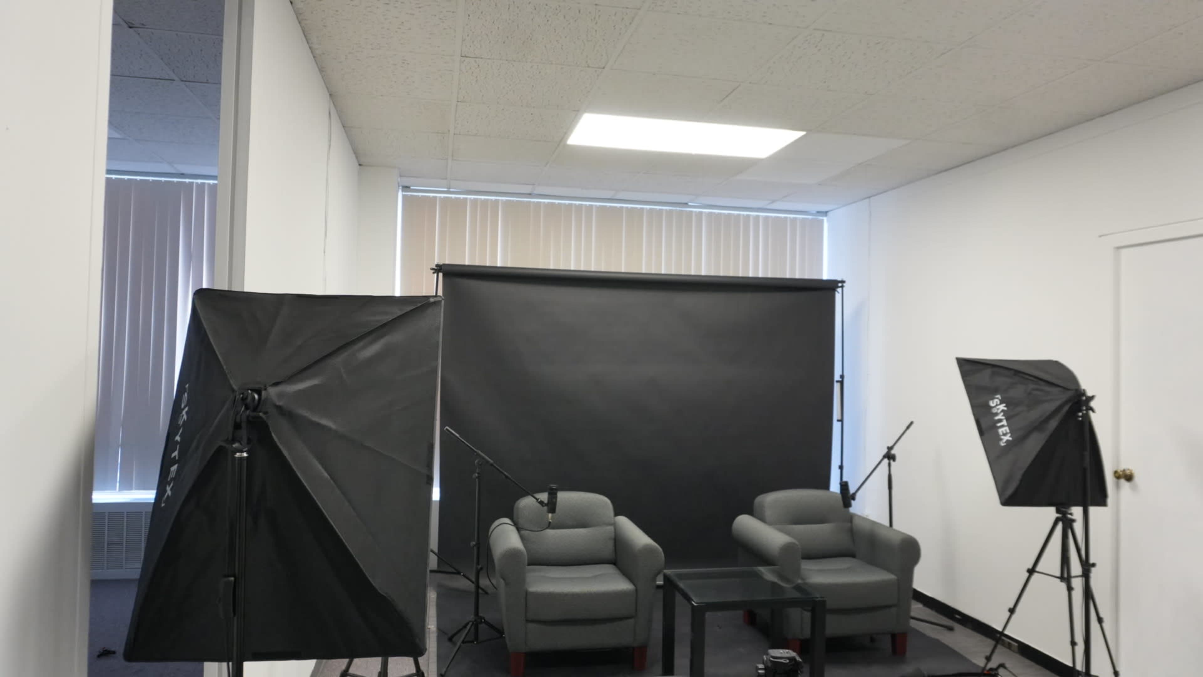 The image shows a studio setup featuring two gray chairs in front of a large black backdrop, with three softbox lights positioned around the area.