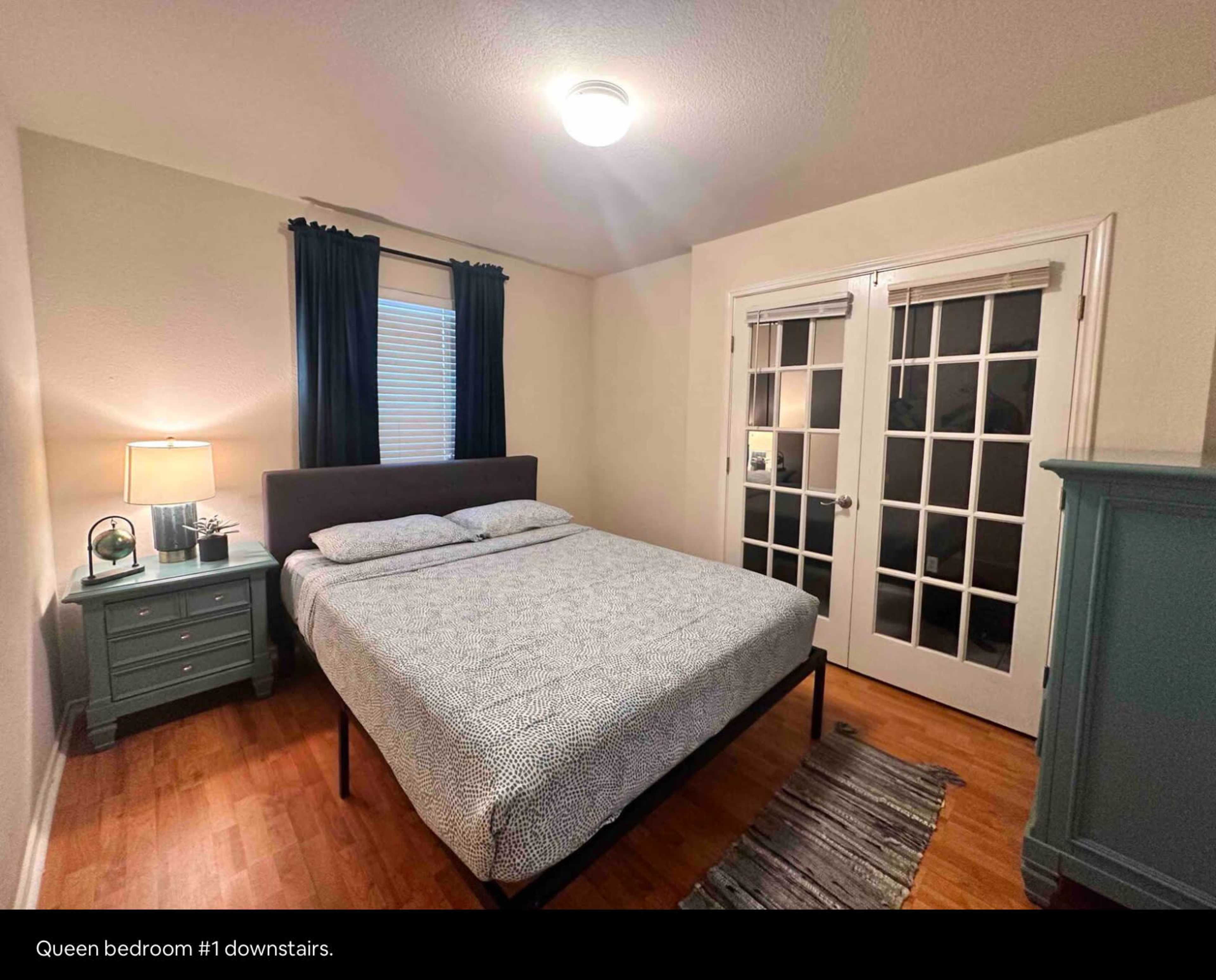 A queen bedroom with a bed, nightstand, lamp, and a set of French doors, all situated on wooden flooring.