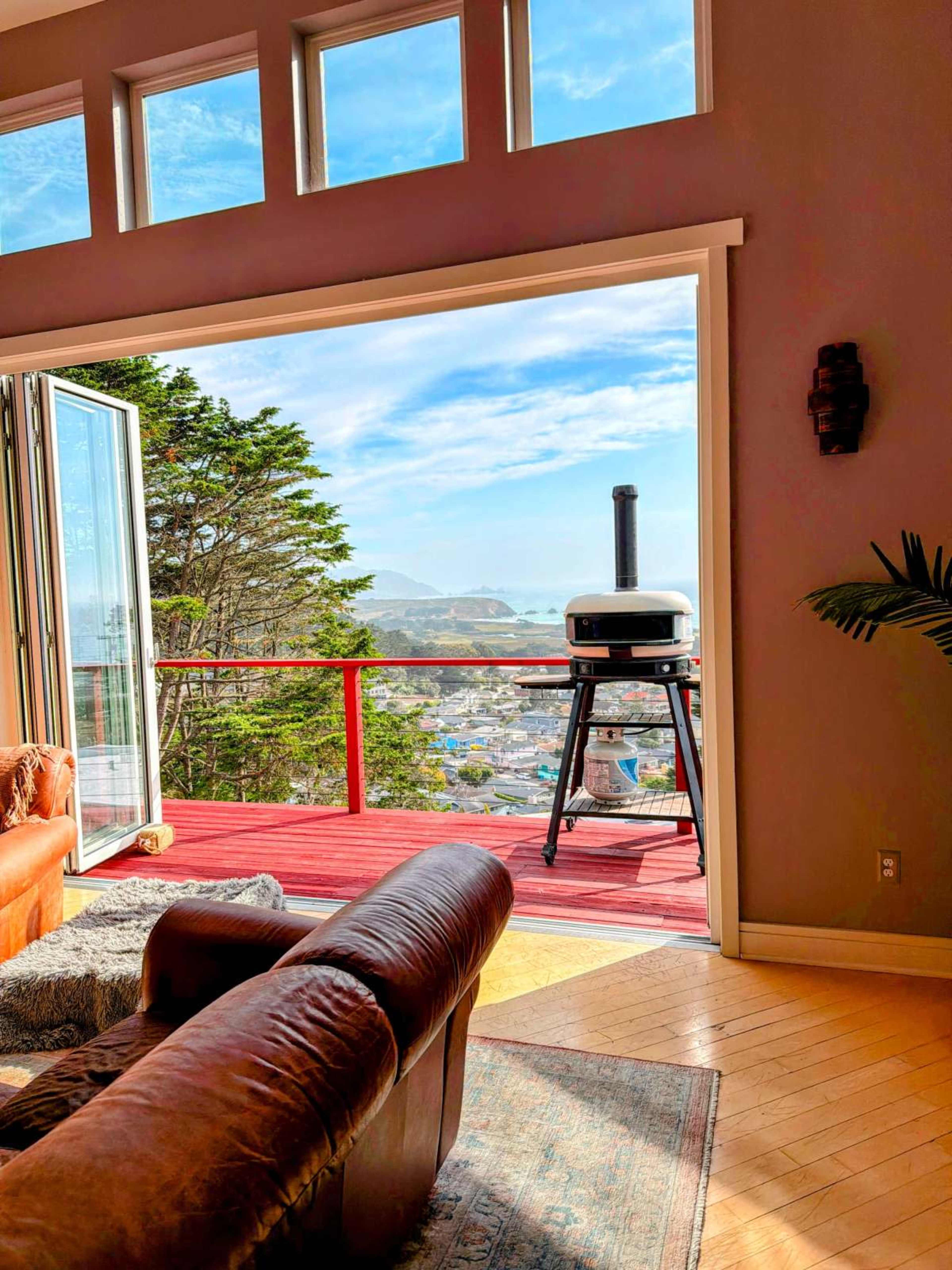 A spacious living room features a leather sofa and opens to a deck with a grill, overlooking a scenic view of hills and a town.