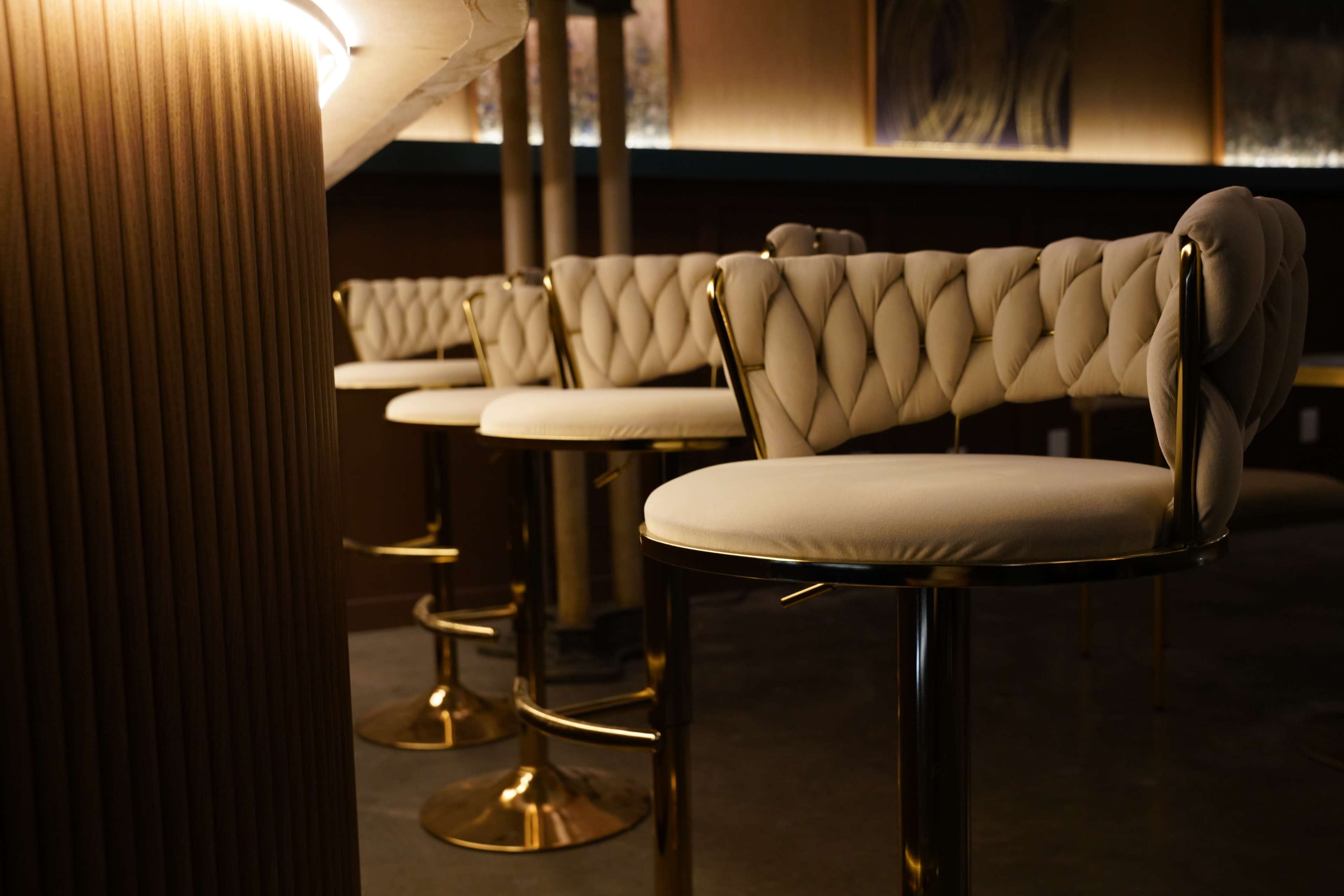 The image shows a row of tufted, high-backed bar stools with gold accents positioned under a softly lit staircase in a modern interior space.