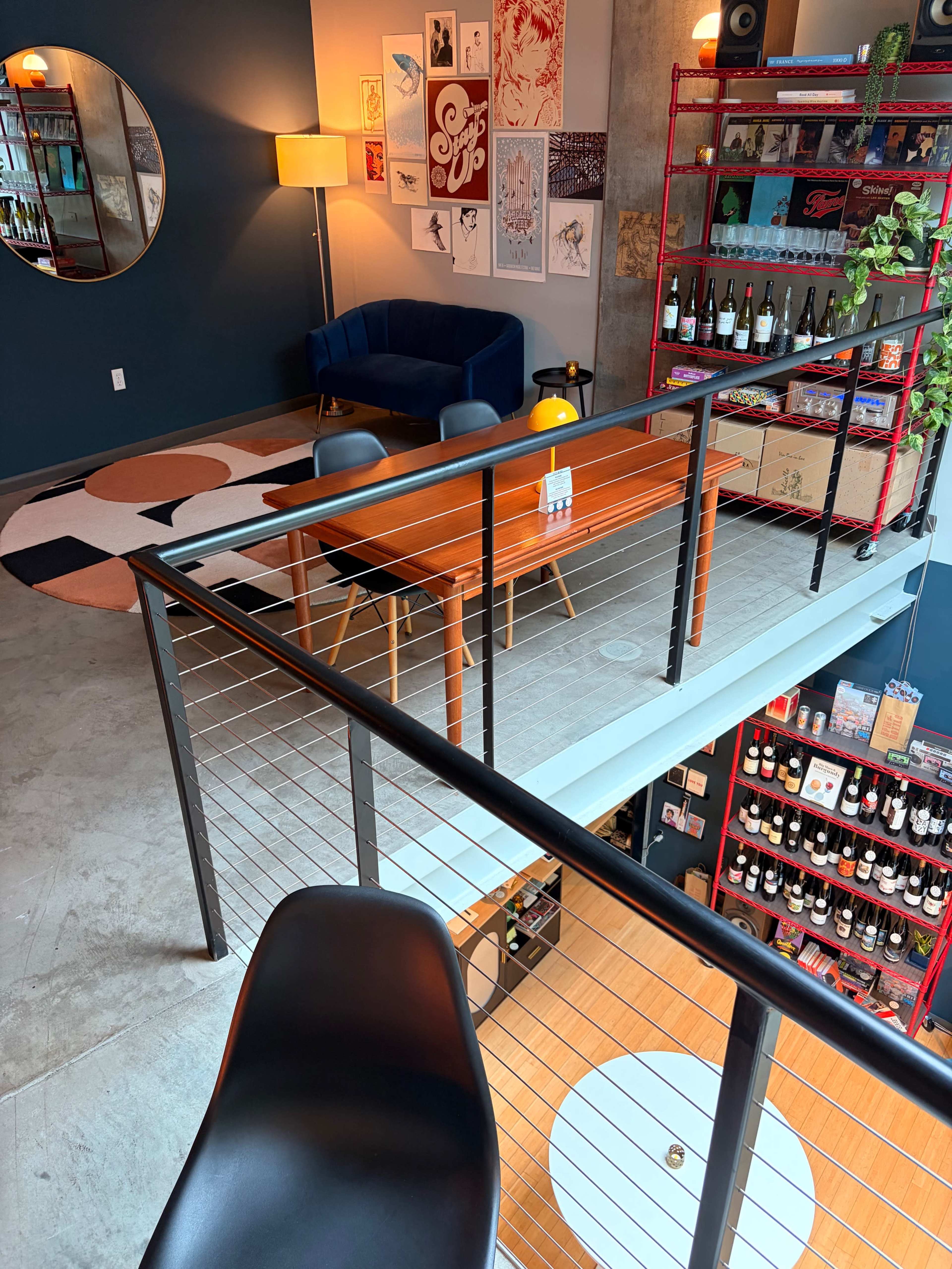 The image shows a multi-level, modern indoor space featuring a seating area with a blue couch, a round rug, and a dining table, along with shelves displaying various wine bottles.
