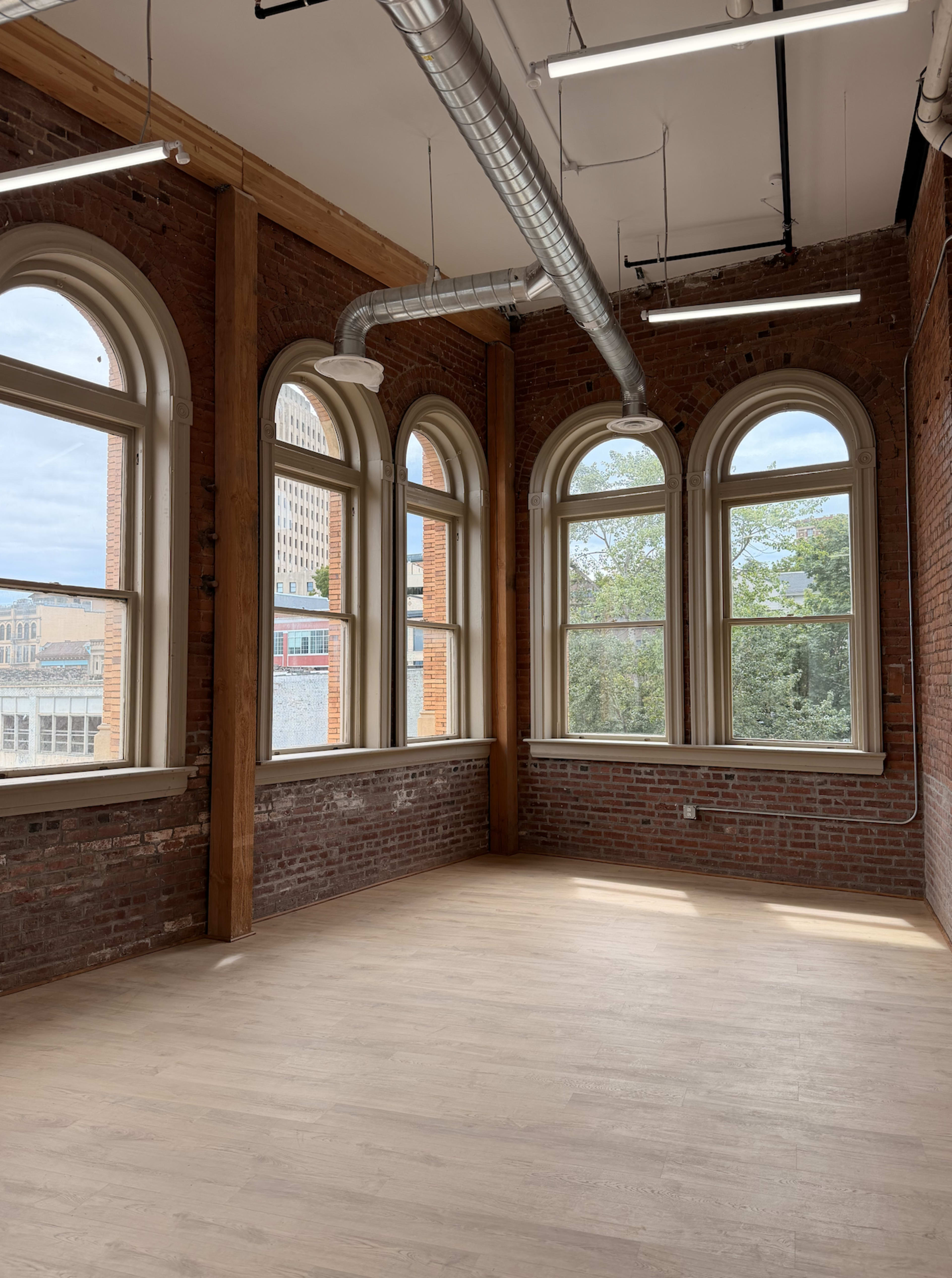 The room features large arched windows, exposed brick walls, and a wooden floor with metal ventilation ducts along the ceiling.