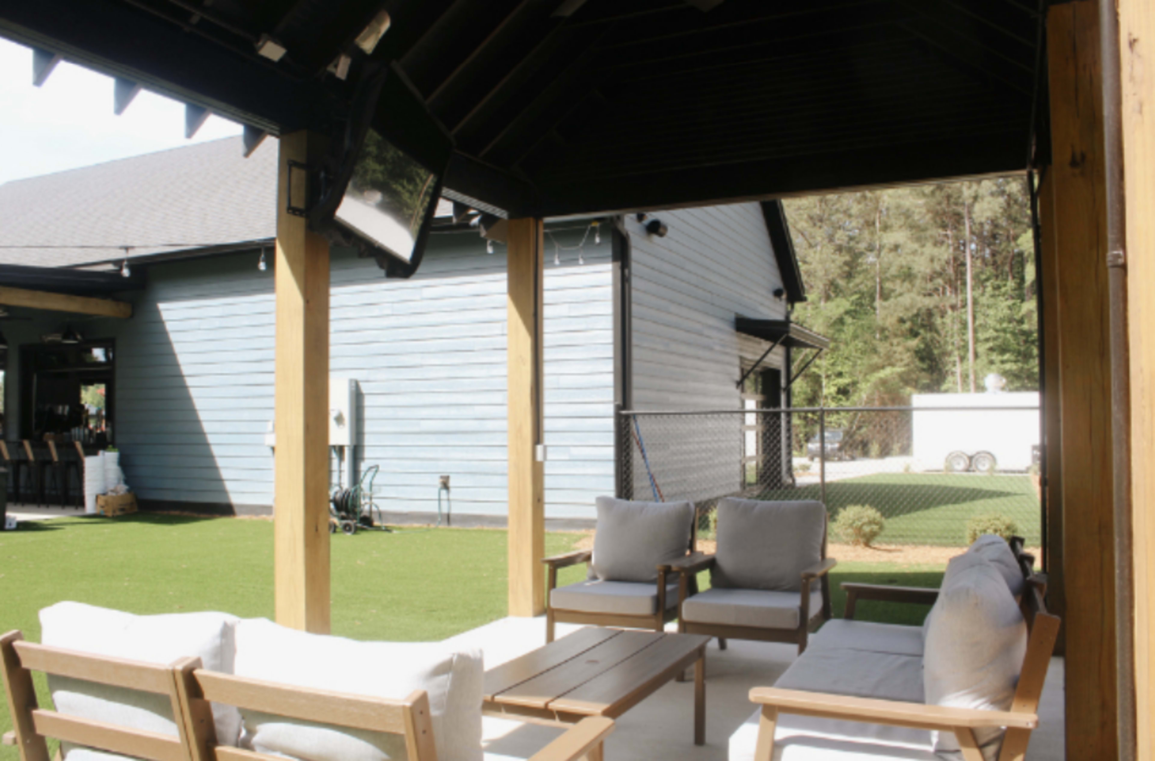 The image shows a covered outdoor seating area with wooden furniture facing a fenced yard behind a blue house.