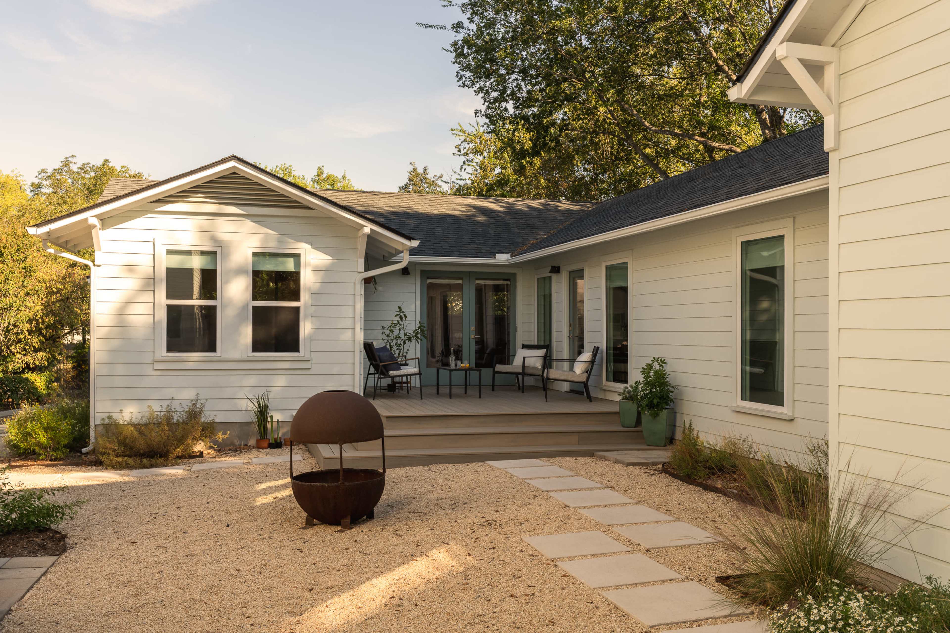 The image shows a modern home with a deck area surrounded by a gravel pathway, plants, and outdoor seating.