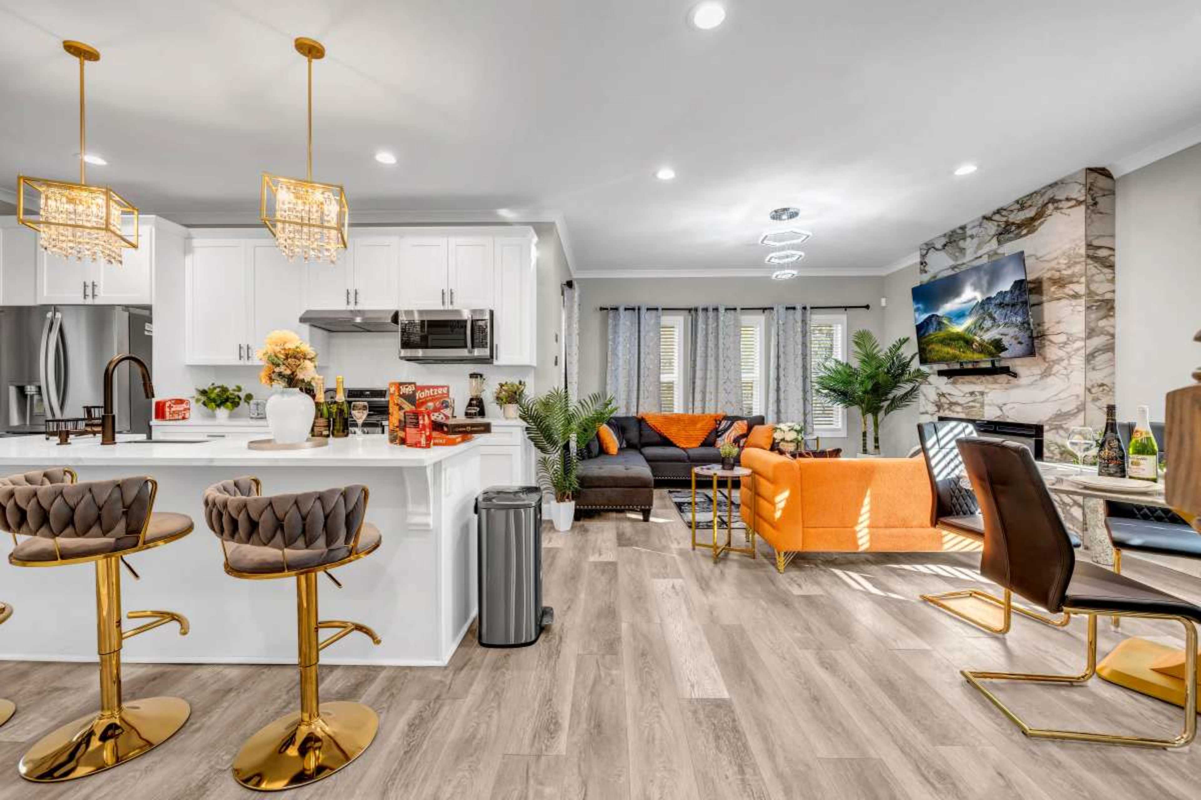 The image shows a modern open-concept living space with a kitchen featuring bar stools, a living area with an orange sofa, and decorative plants.