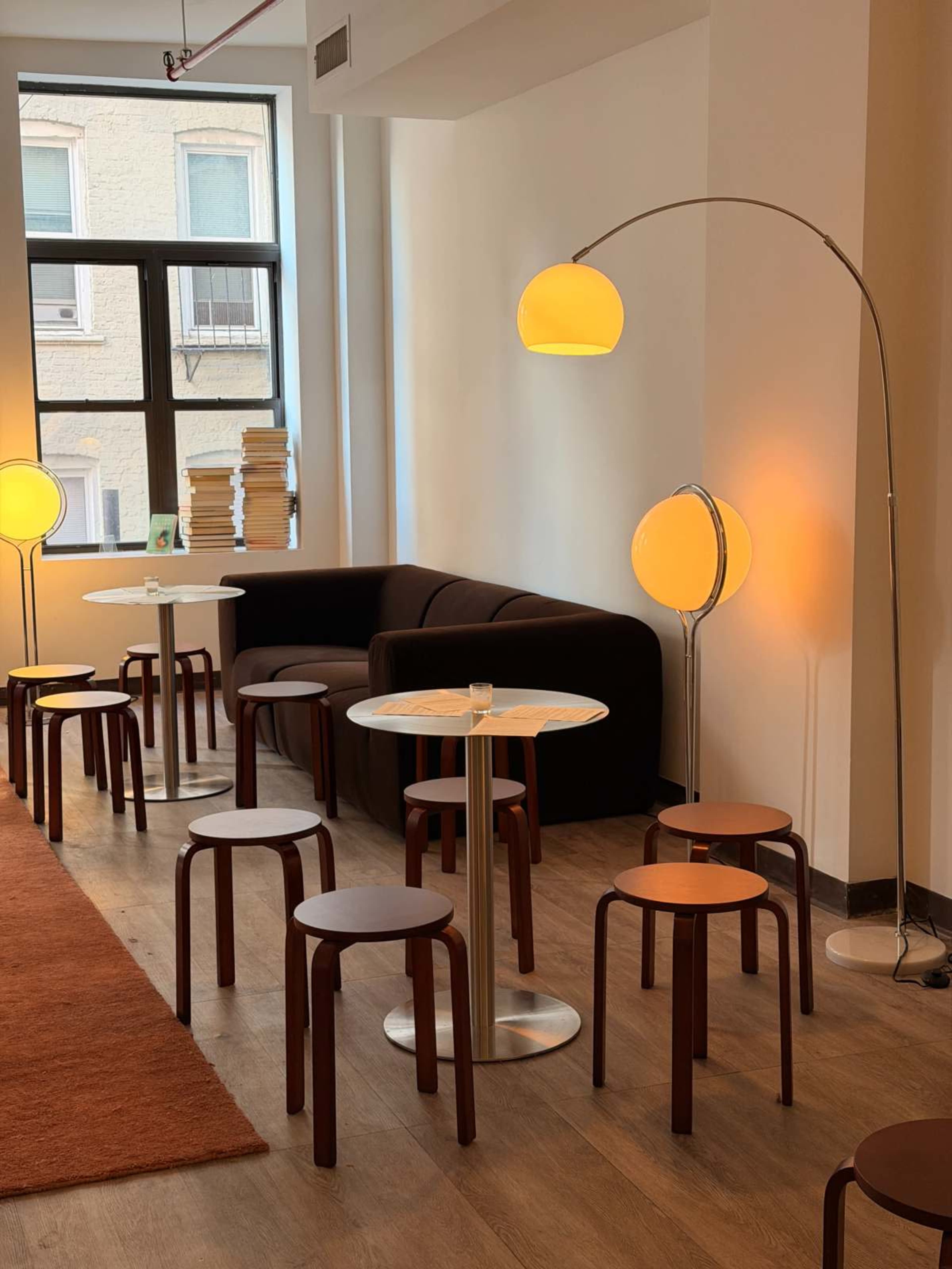 The image shows a cozy café interior featuring a dark brown sofa, several small tables with stools, and two distinct floor lamps casting a warm glow.