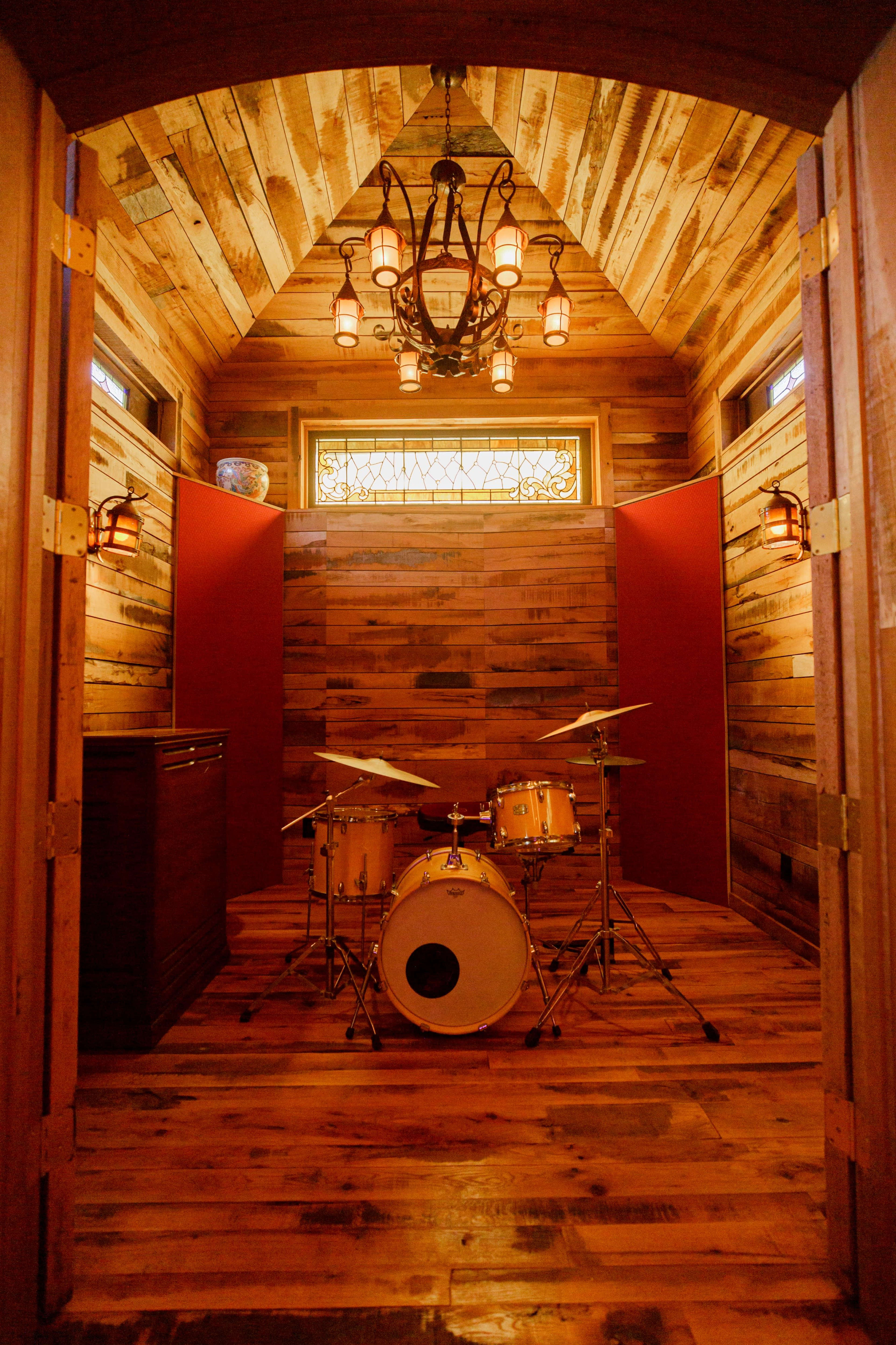 A wooden-paneled music room features a drum set at the center, illuminated by a chandelier overhead.