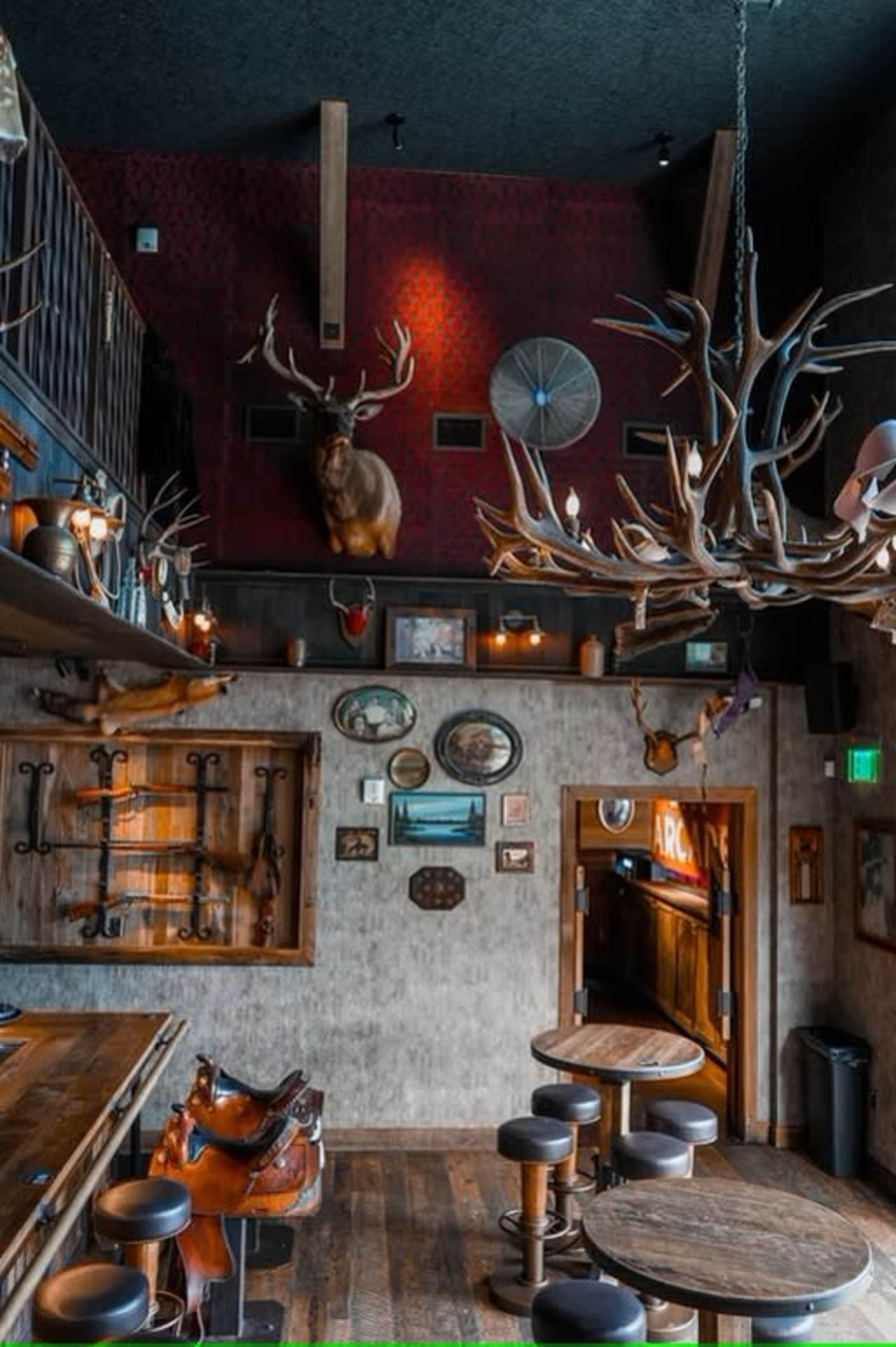 The image depicts the interior of a rustic bar featuring wooden furniture, mounted deer antlers on the walls, and a staircase leading to an upper level.