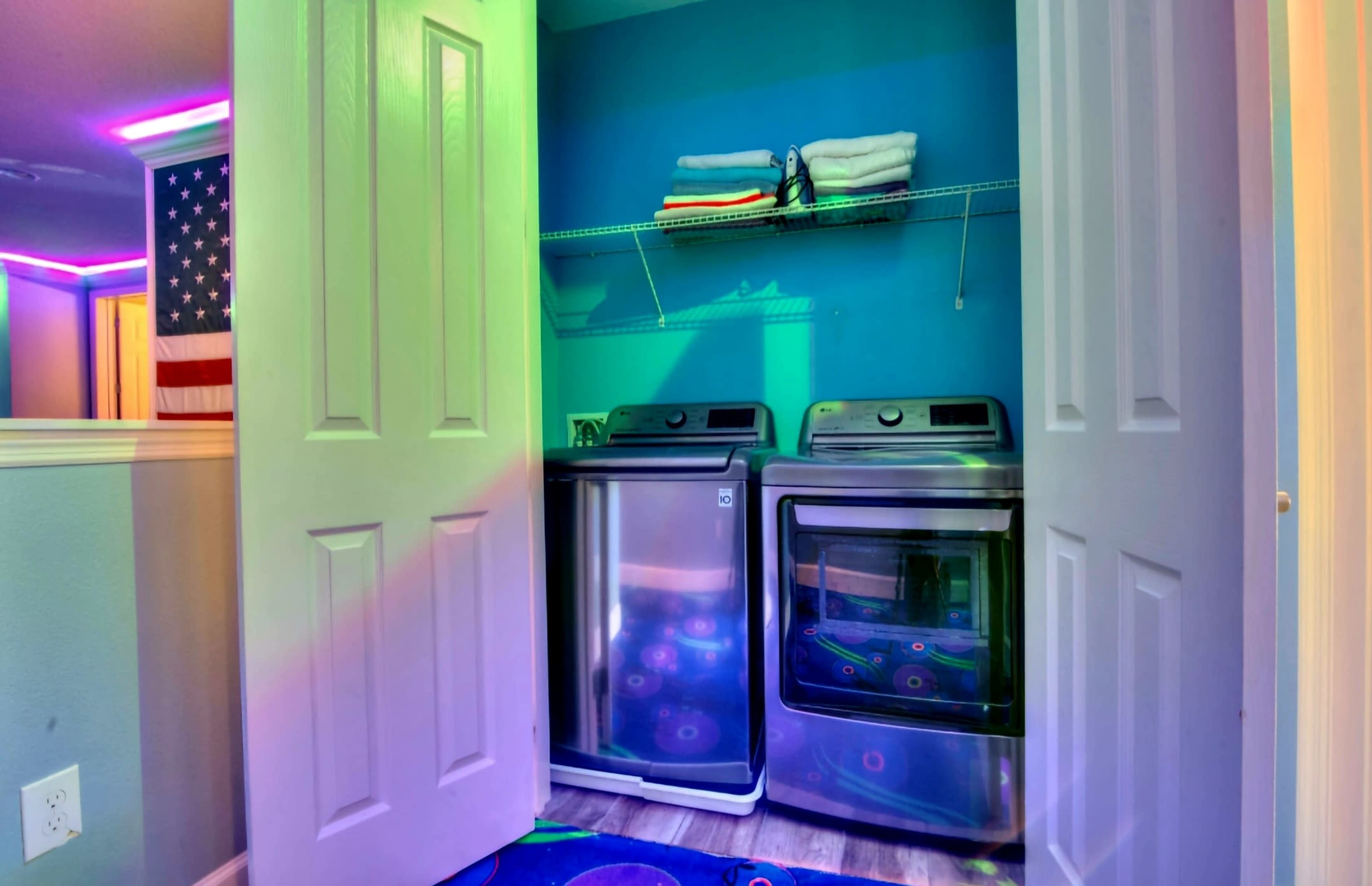 A laundry room features stacked washer and dryer units with shelves above them holding folded towels, set against a blue wall.