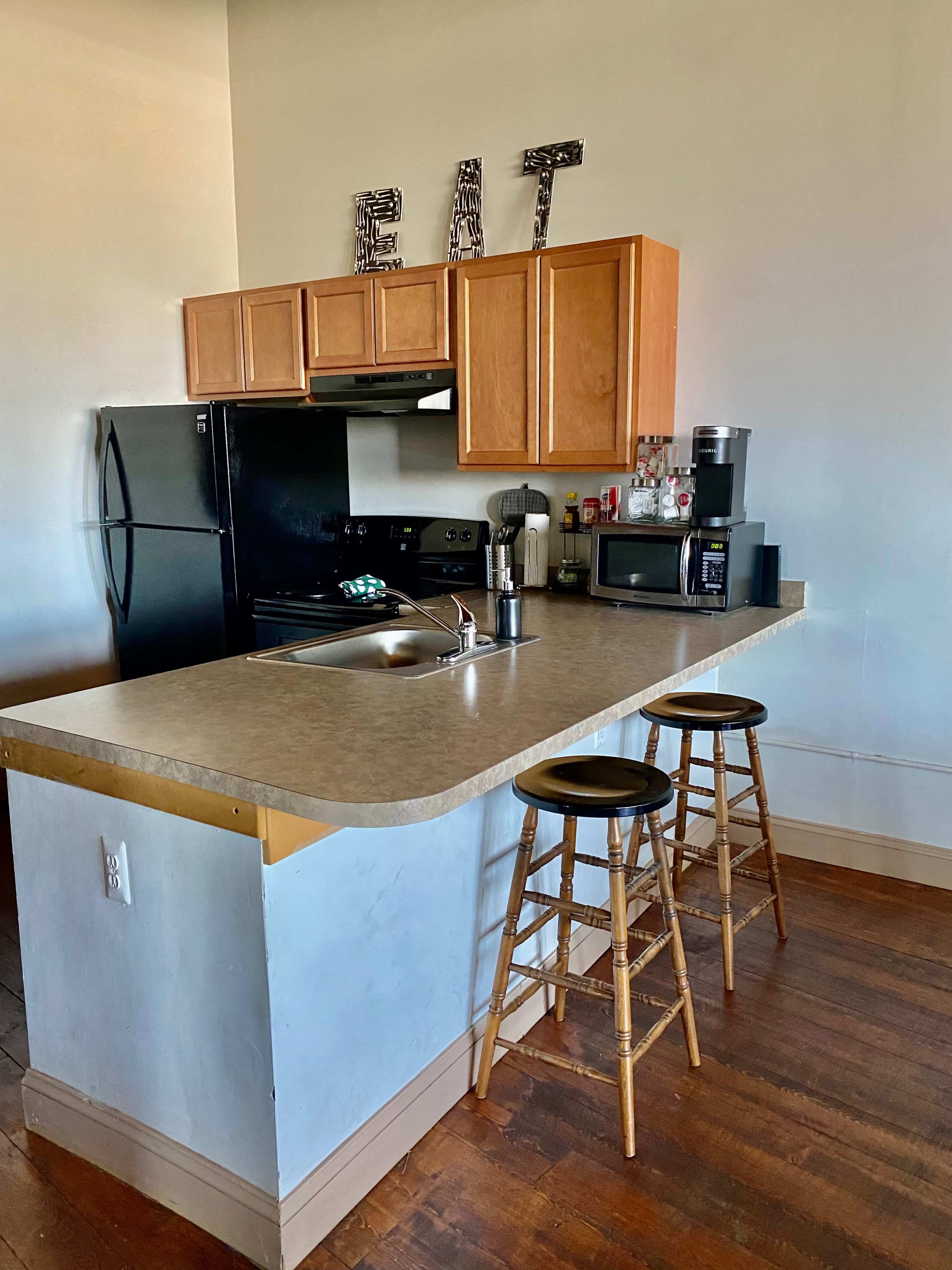 A kitchen features wooden cabinets, a black refrigerator, a stove, a microwave, and two bar stools at a counter.