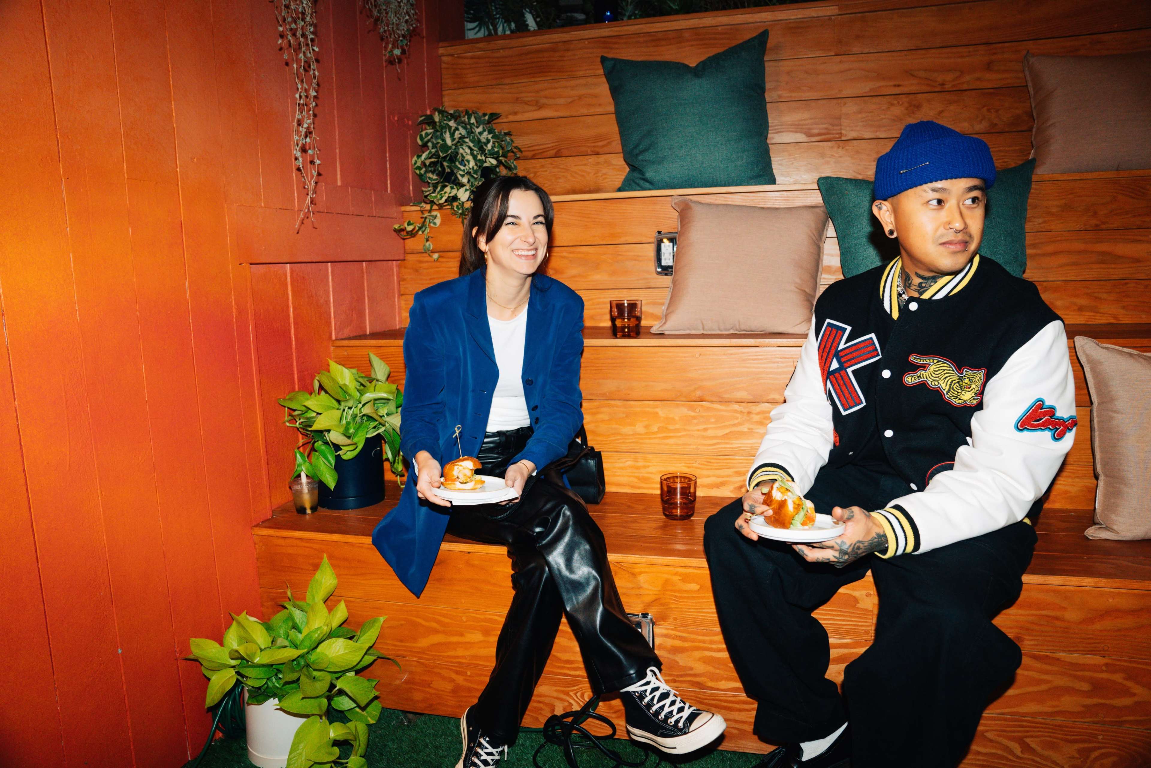 A woman and a man sit on a tiered wooden bench holding plates of food in a brightly colored setting.