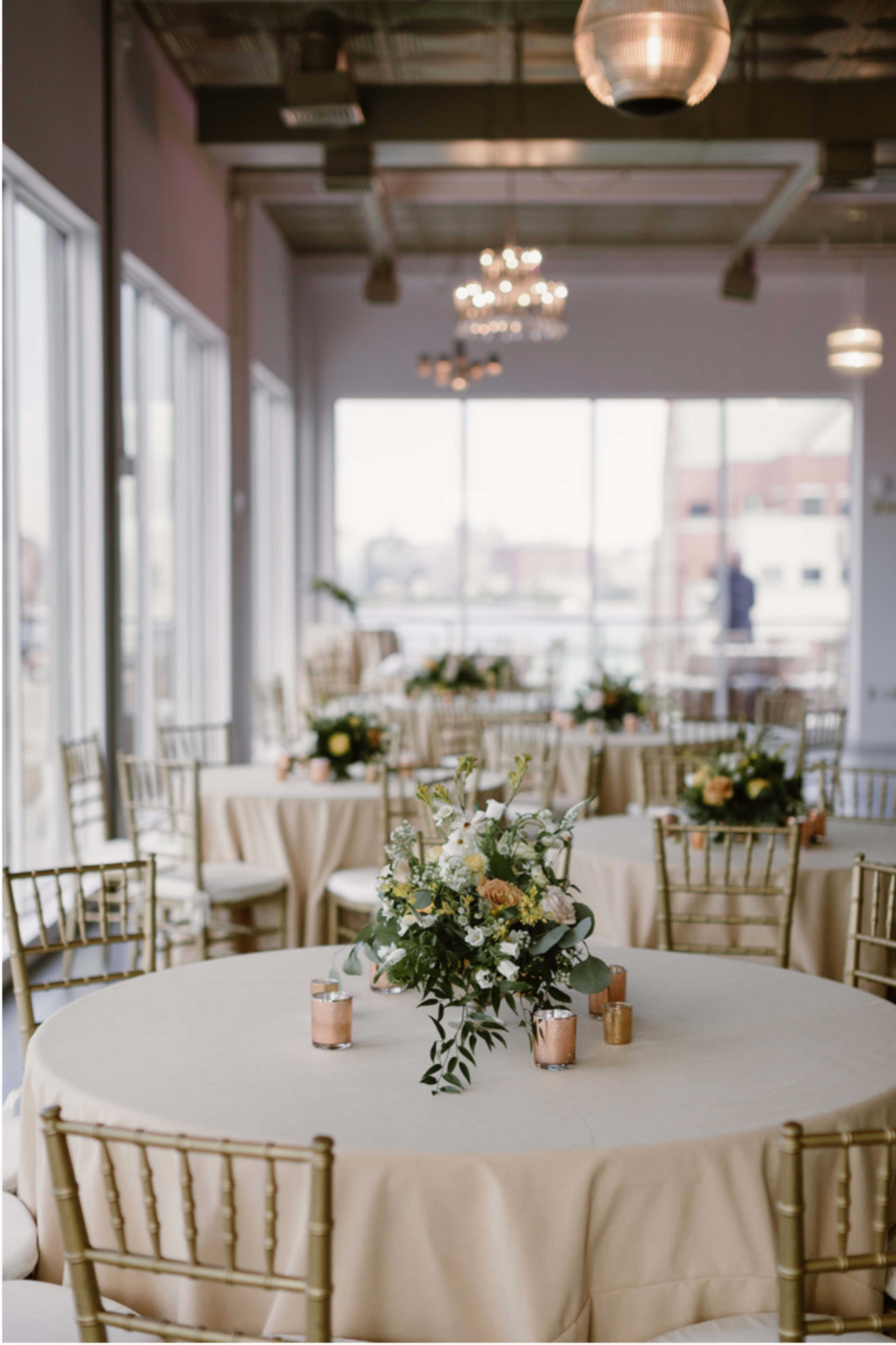 A dining area with round tables covered in beige linens, each adorned with floral centerpieces and small candles, set within a venue with large windows.