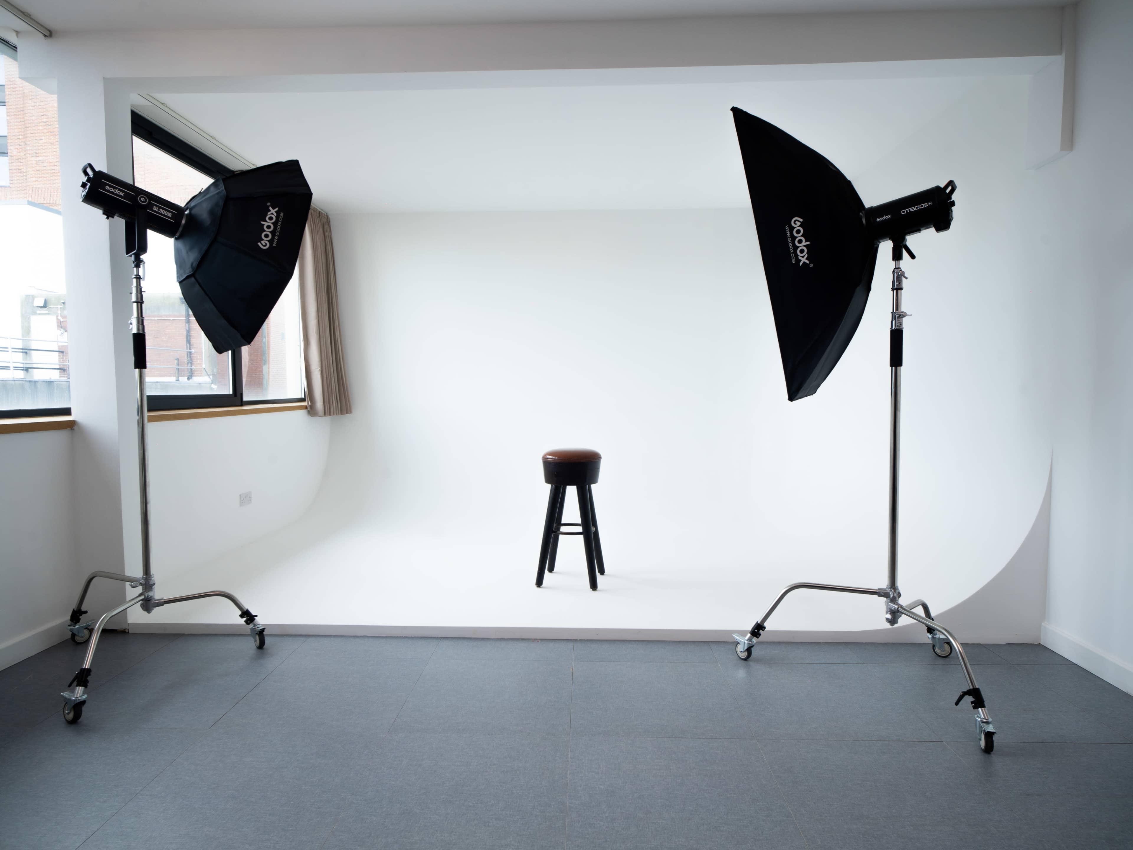 The image shows a photography studio with a white backdrop, a stool in the center, and two light setups on either side.