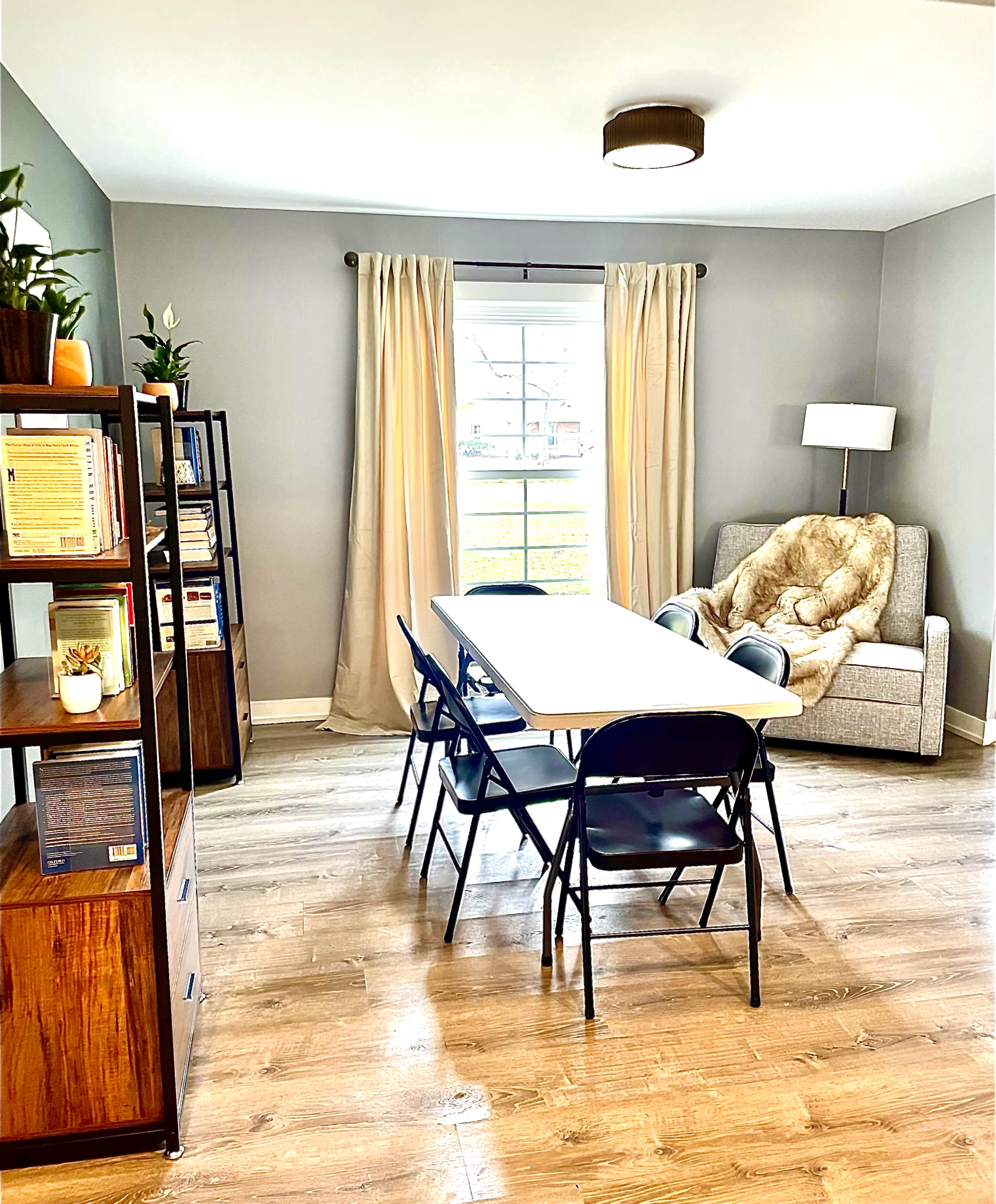 A dining area features a long table surrounded by black chairs, with a bookshelf and a cozy seating area next to a window.