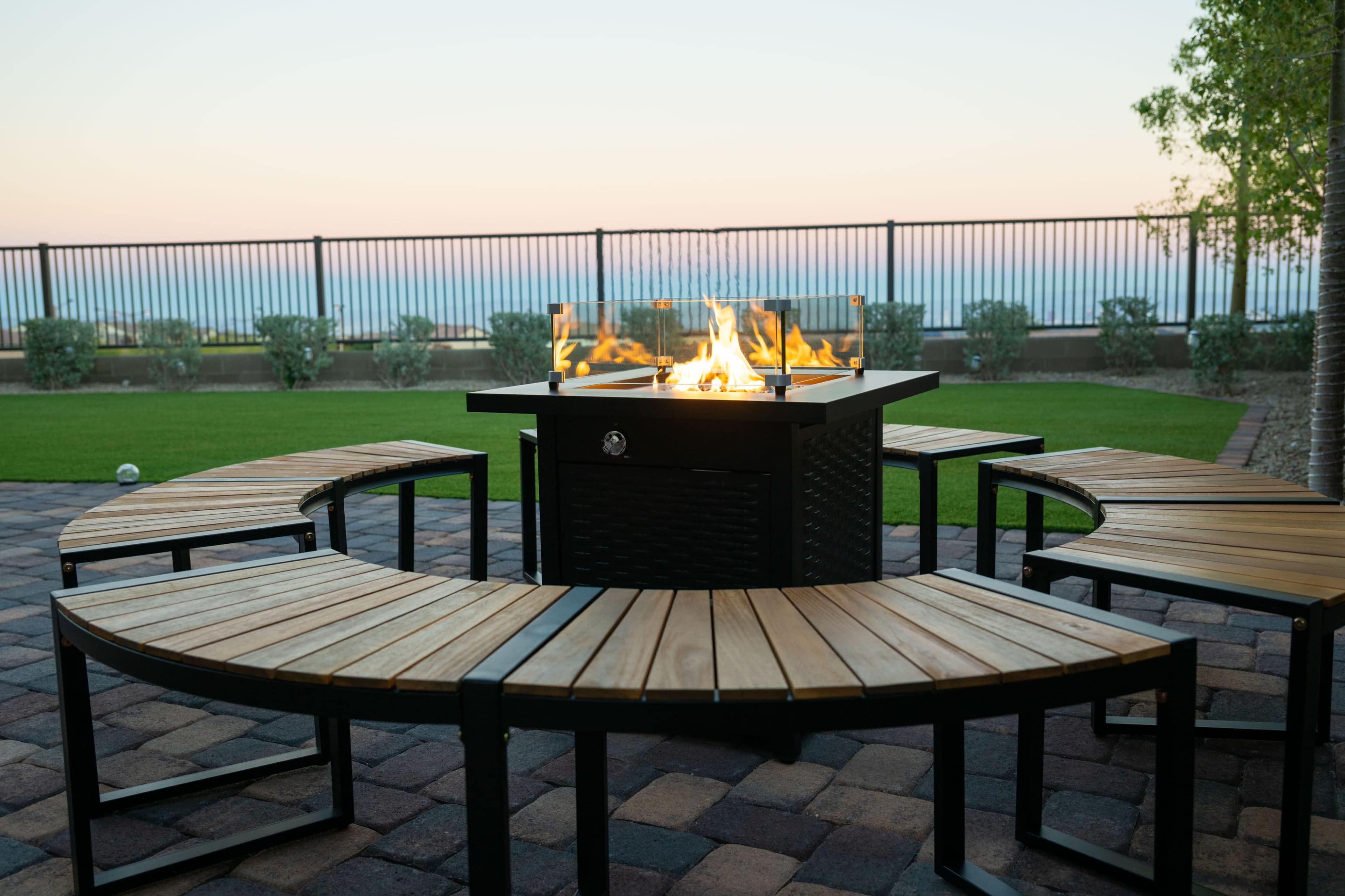 A circular arrangement of wooden benches surrounds a fire pit on a patio overlooking a landscape at dusk.