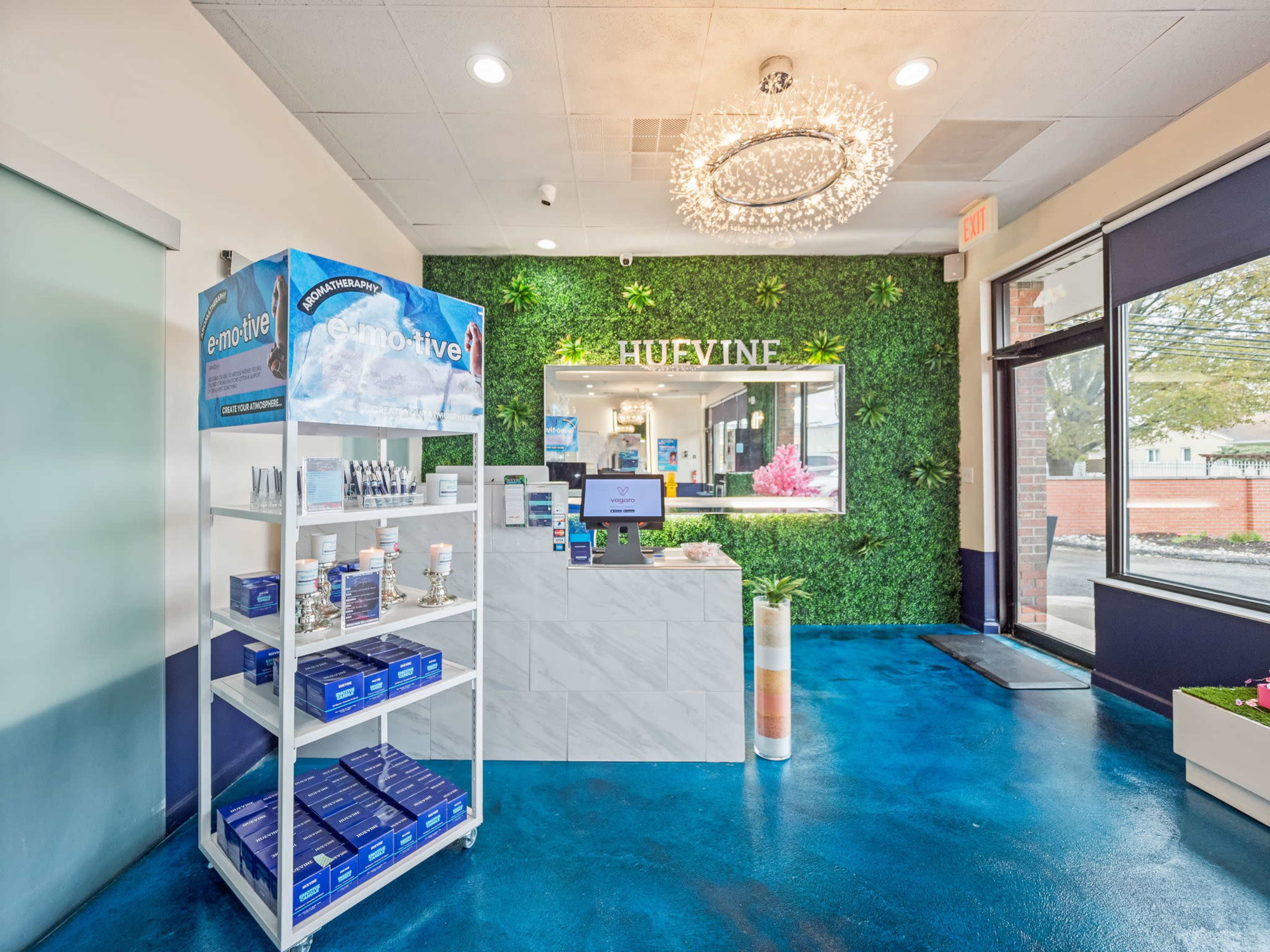 An indoor reception area of a business featuring a green wall, a display stand with products, a counter with digital screens, and large windows allowing natural light.