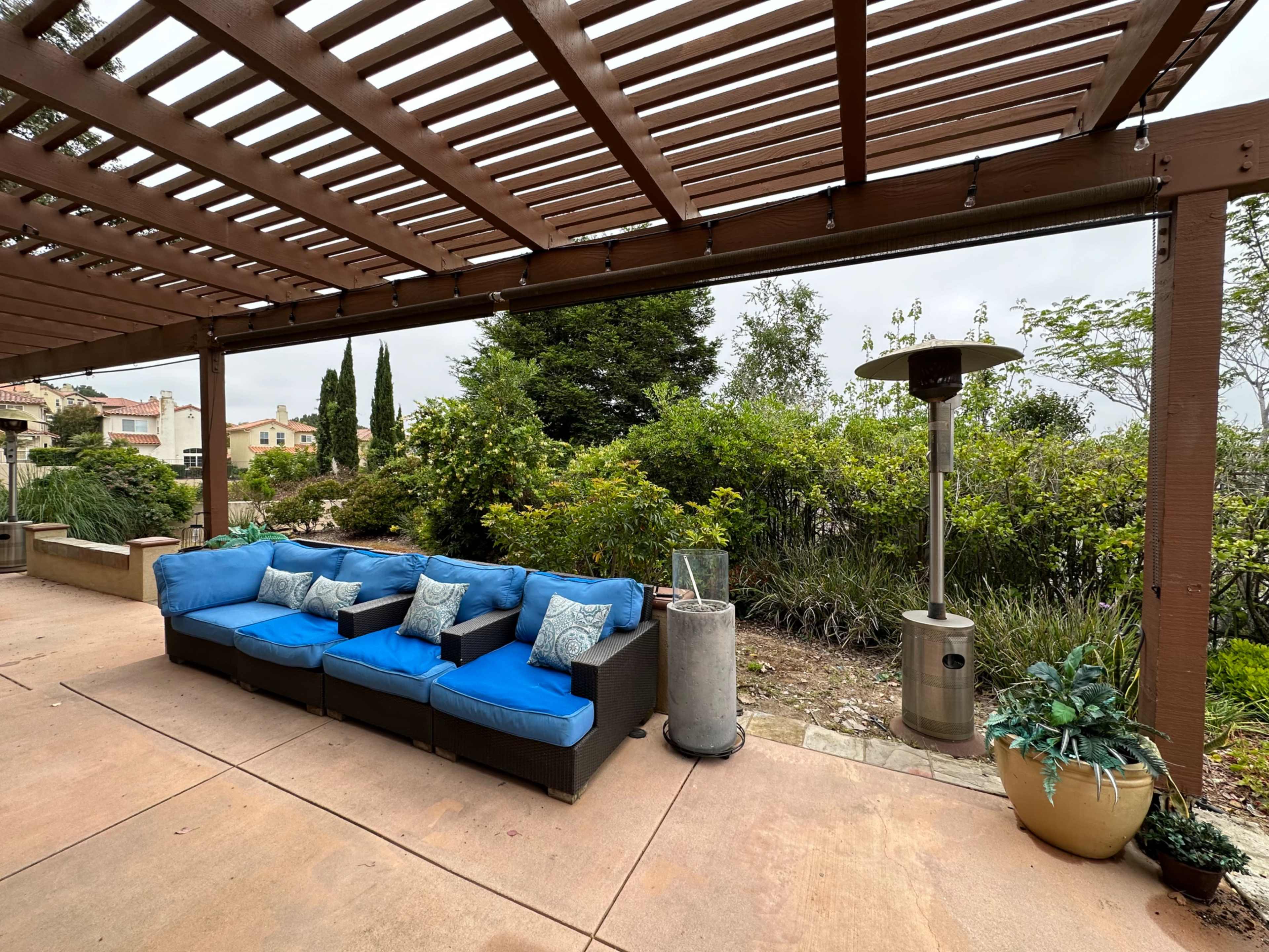 A blue outdoor sectional sofa is placed under a wooden pergola surrounded by greenery and landscaping features.