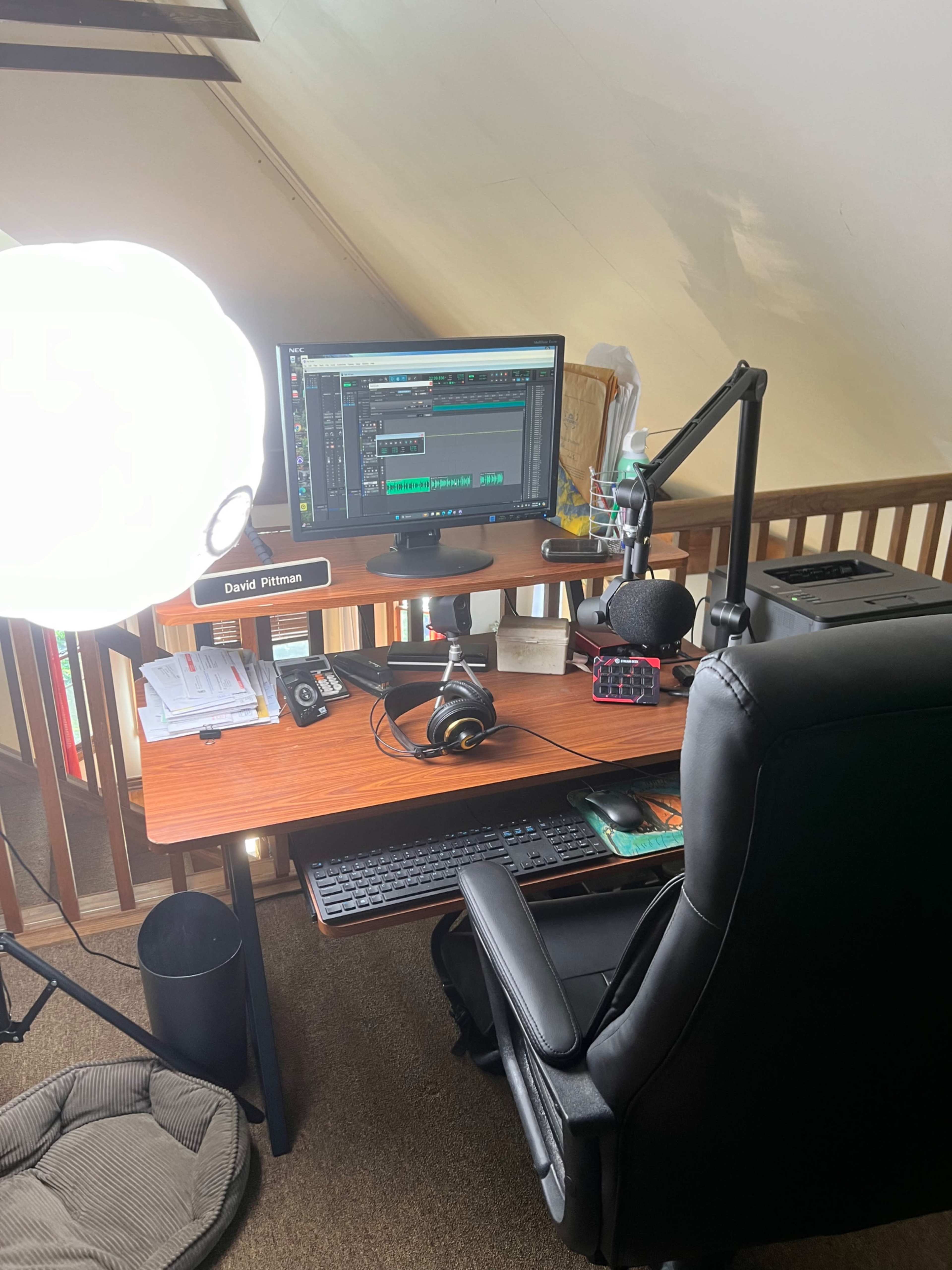A wooden desk is set up in a loft space with a computer monitor displaying audio editing software, a microphone on a stand, headphones, and a large light source nearby.