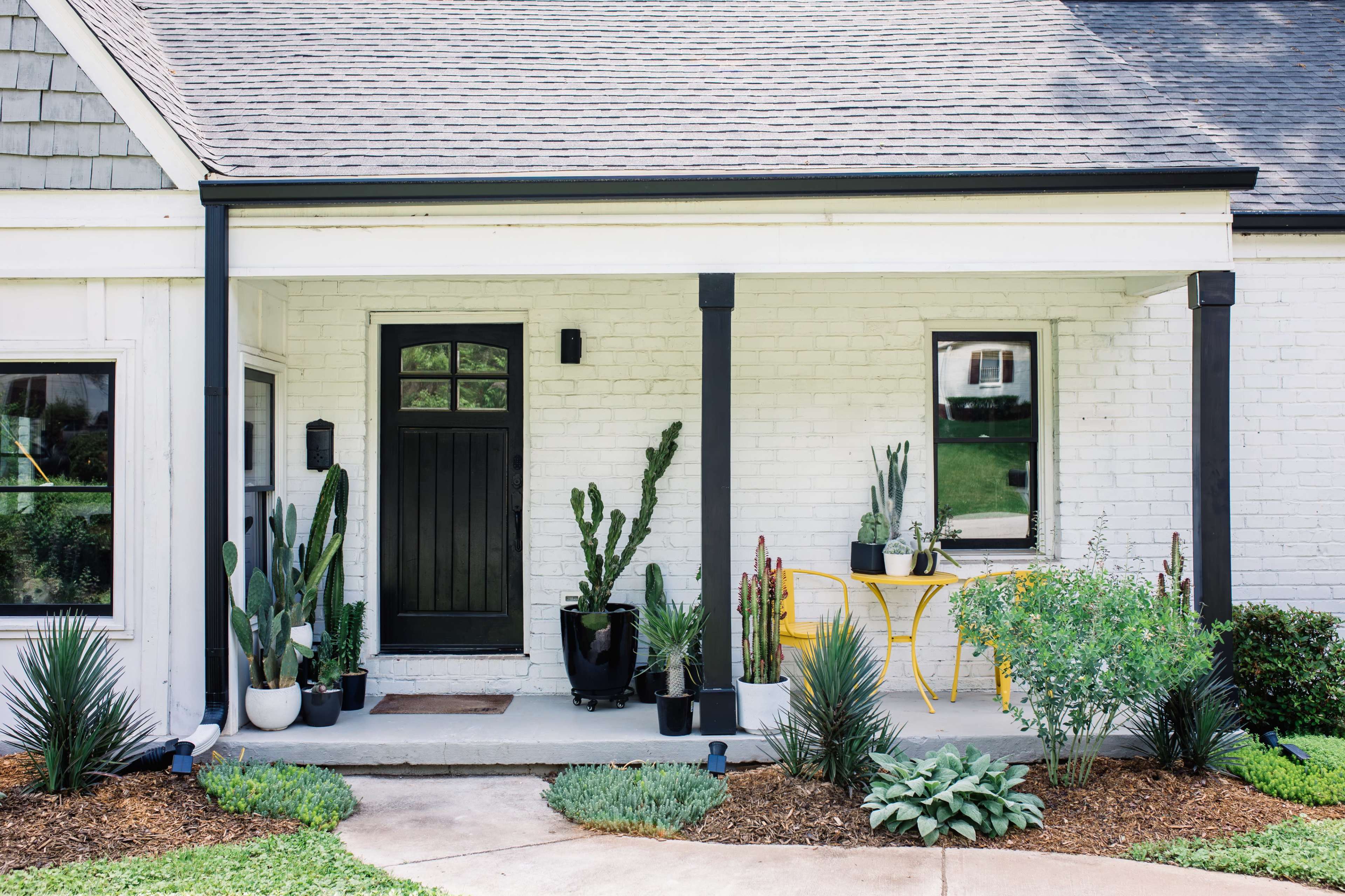 The image shows a front porch of a house with a black door, several potted plants, and a yellow table beside a garden bed.
