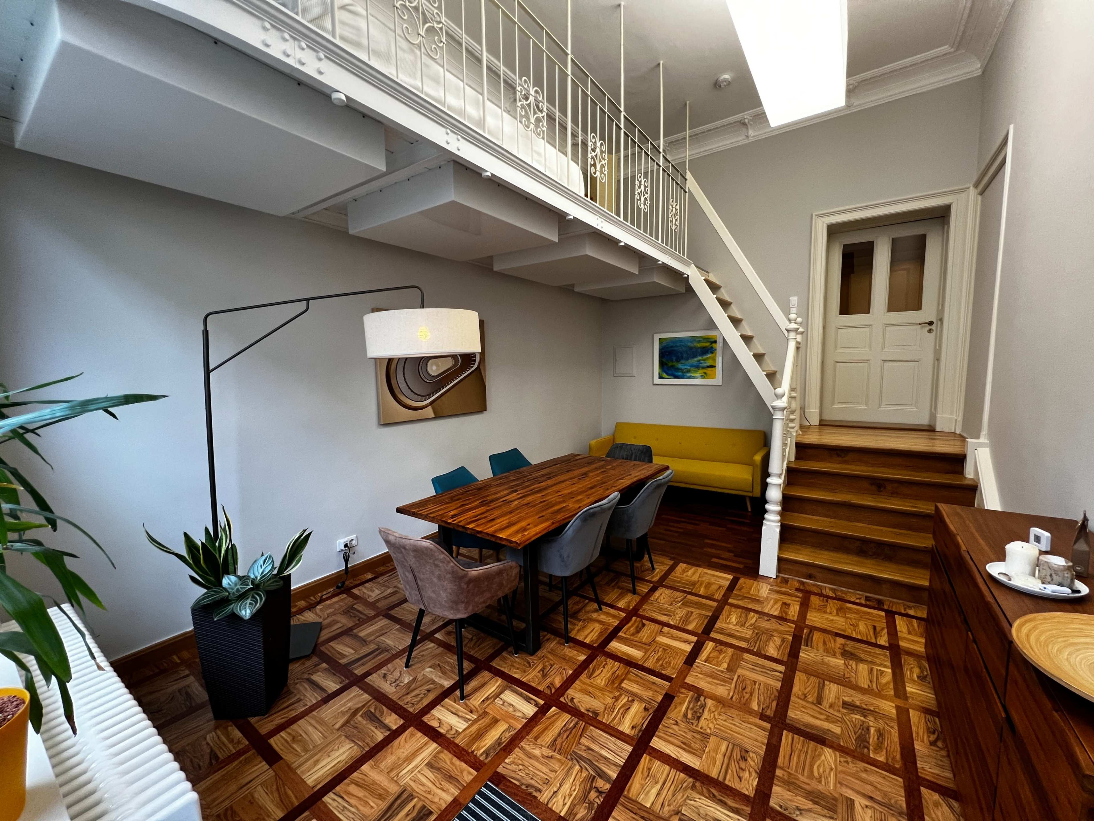 The image shows a modern dining area with a wooden table surrounded by chairs, a floor lamp, and stairs leading to an upper level.