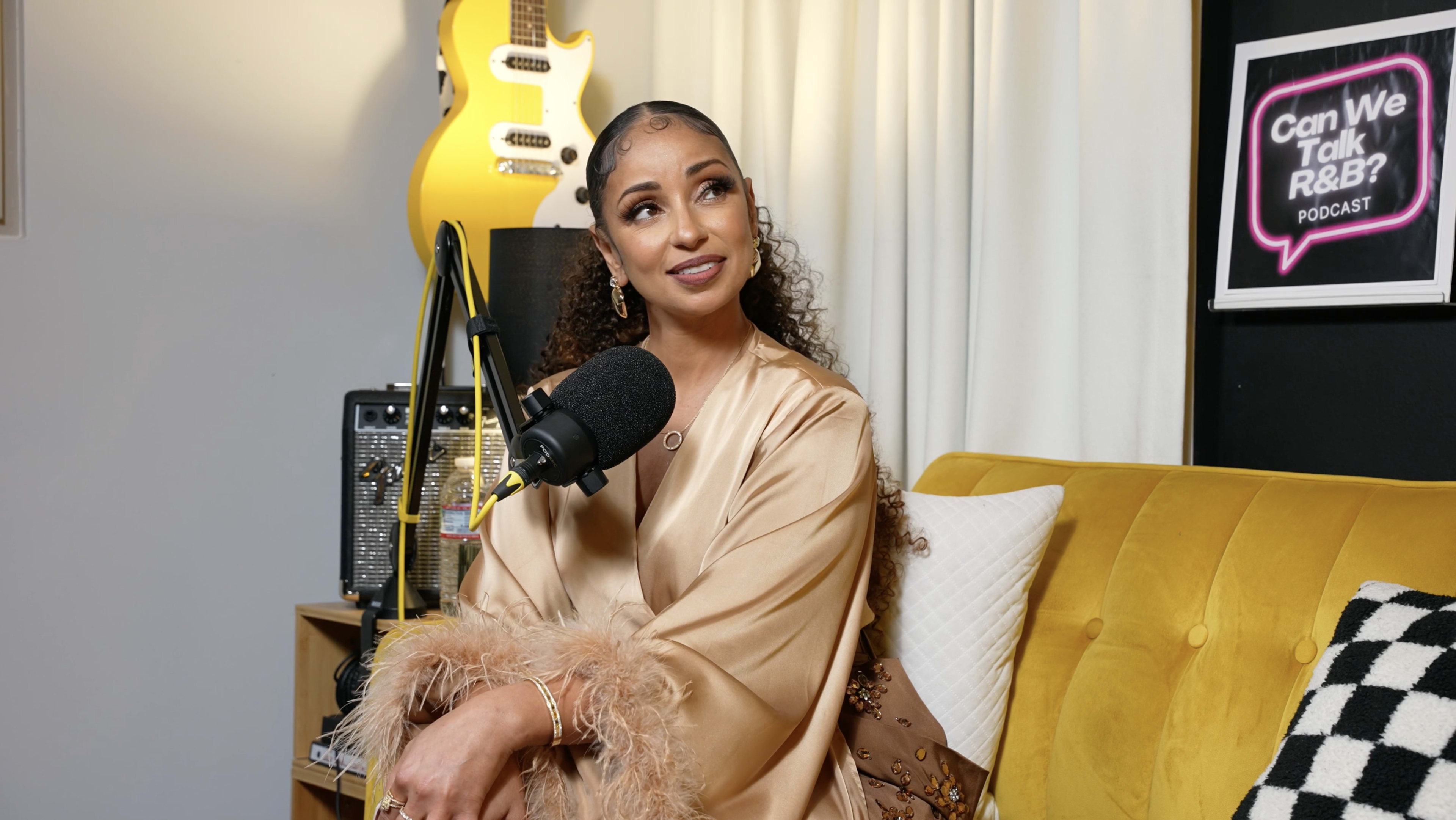 A woman with curly hair sits on a yellow couch in a podcast studio, smiling while speaking into a microphone.