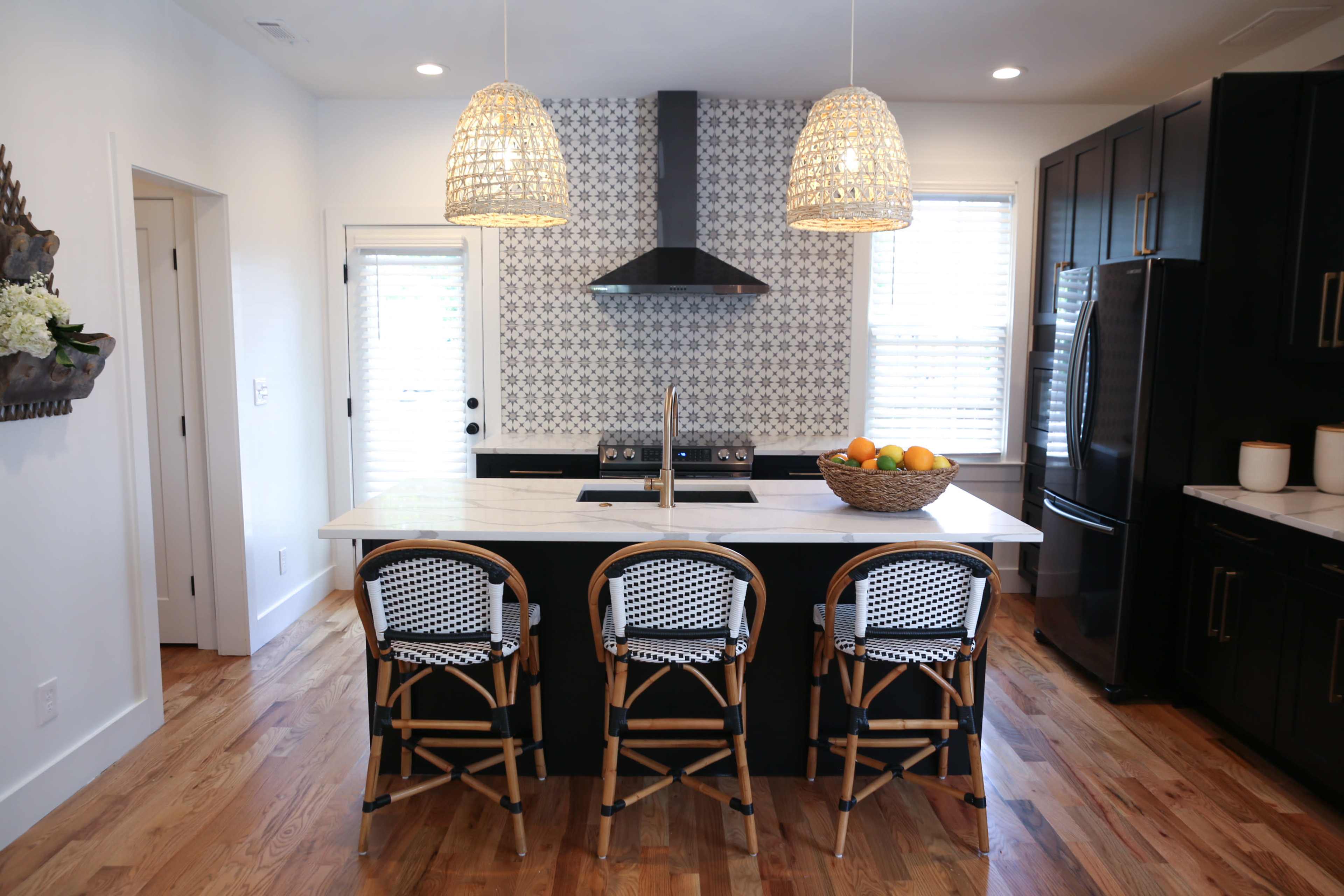 A modern kitchen features a central island with three patterned stools, black cabinetry, a stainless steel refrigerator, and pendant lights above.