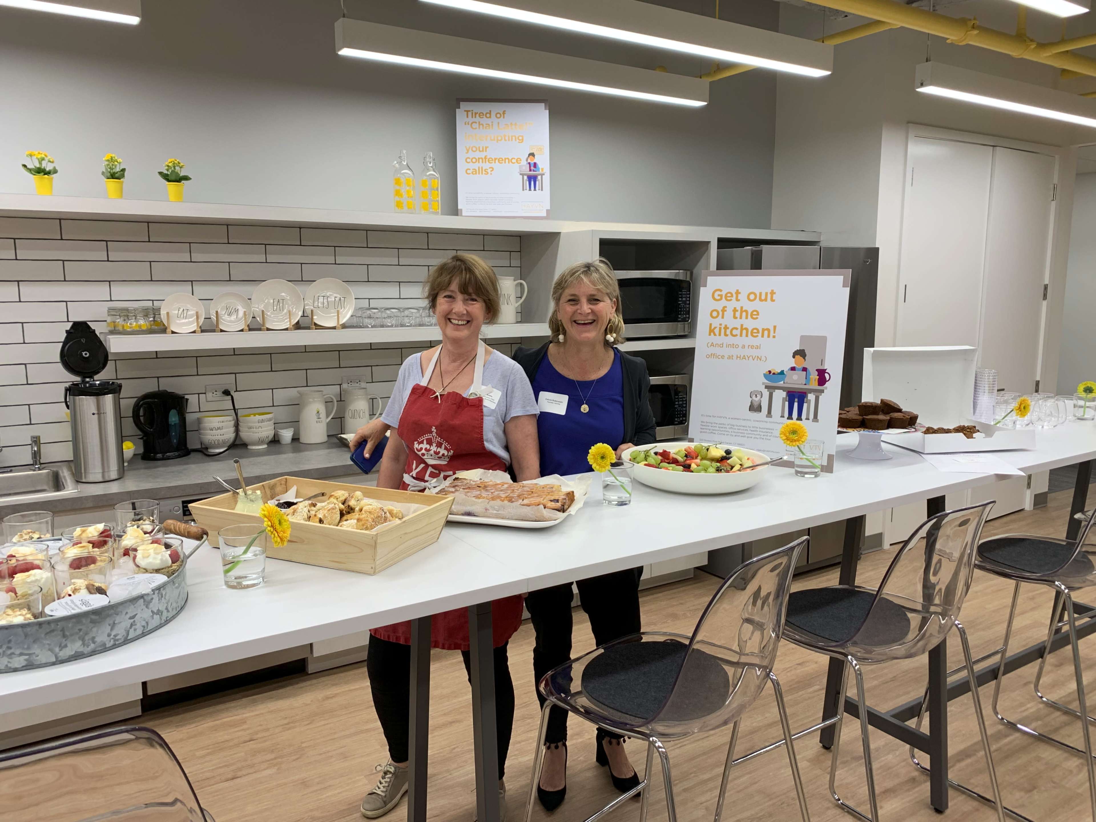 Two women stand in a kitchen area beside a table covered with various desserts and a sign that says "Get out of the kitchen!"