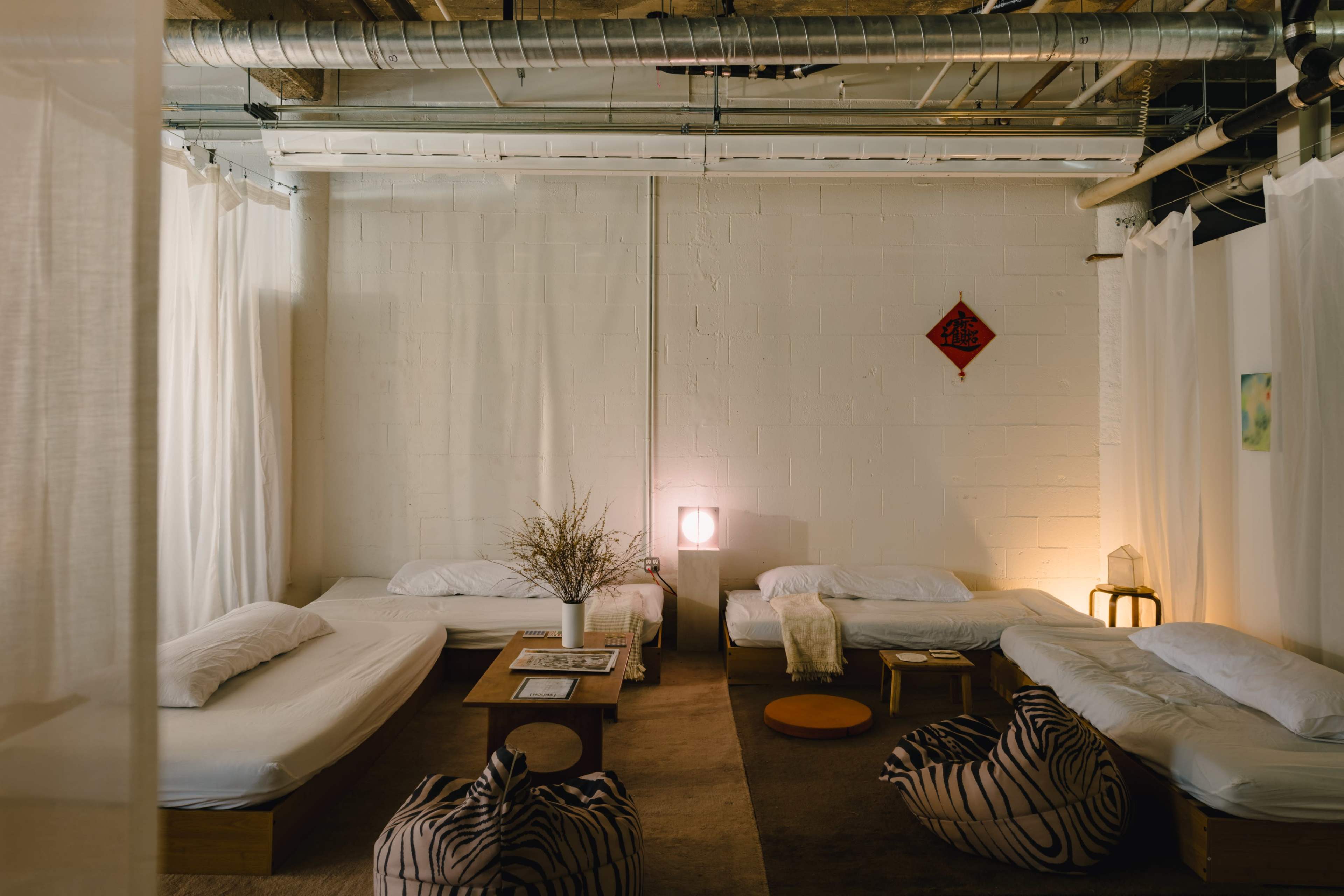 The image shows a minimalist room with several beds arranged around a central wooden table, softly lit by ambient lamps, and separated by sheer curtains.