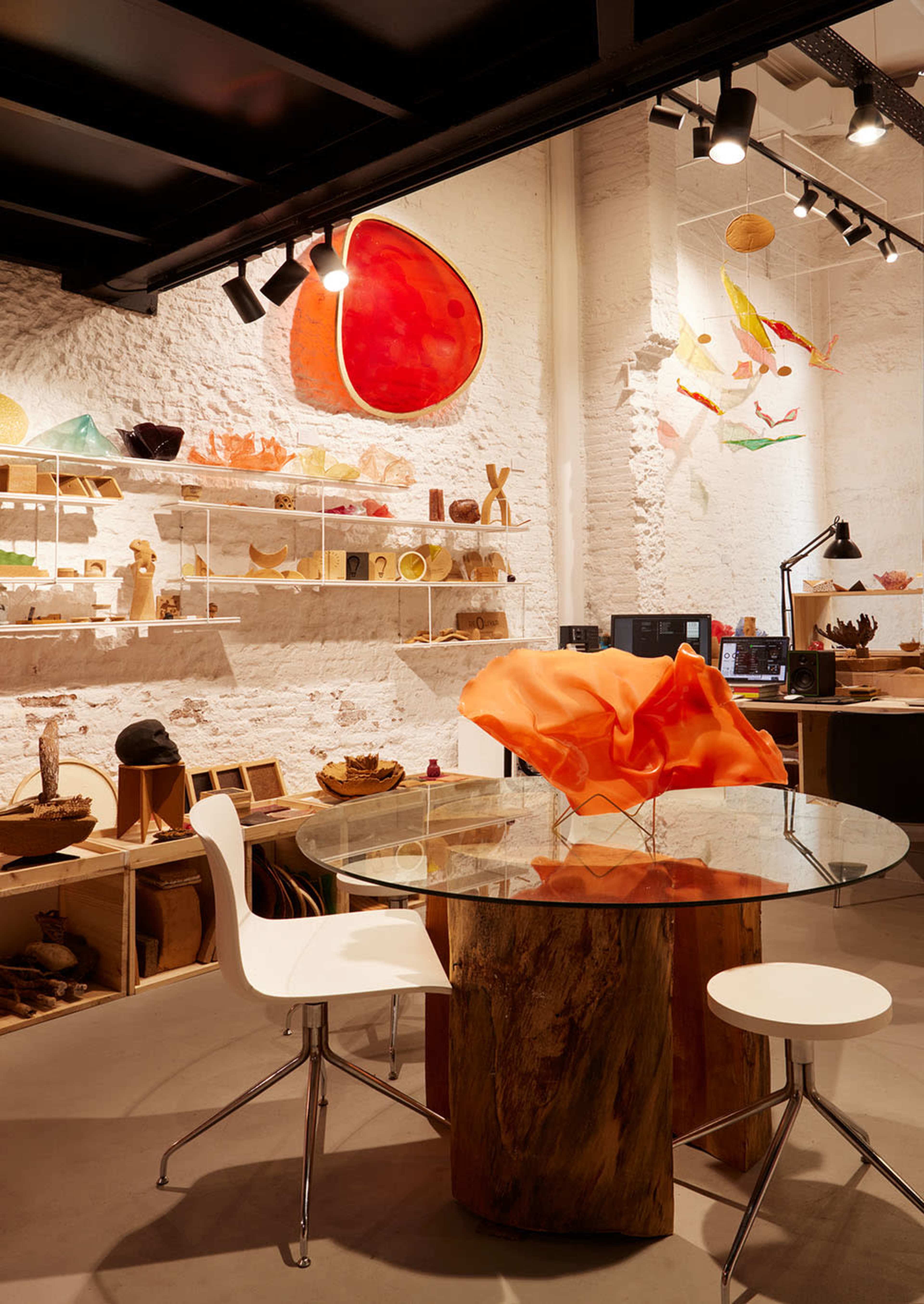 The image shows a modern workspace featuring a round glass table with wooden and white chairs, surrounded by colorful artistic installations on the walls and shelves filled with various crafted items.