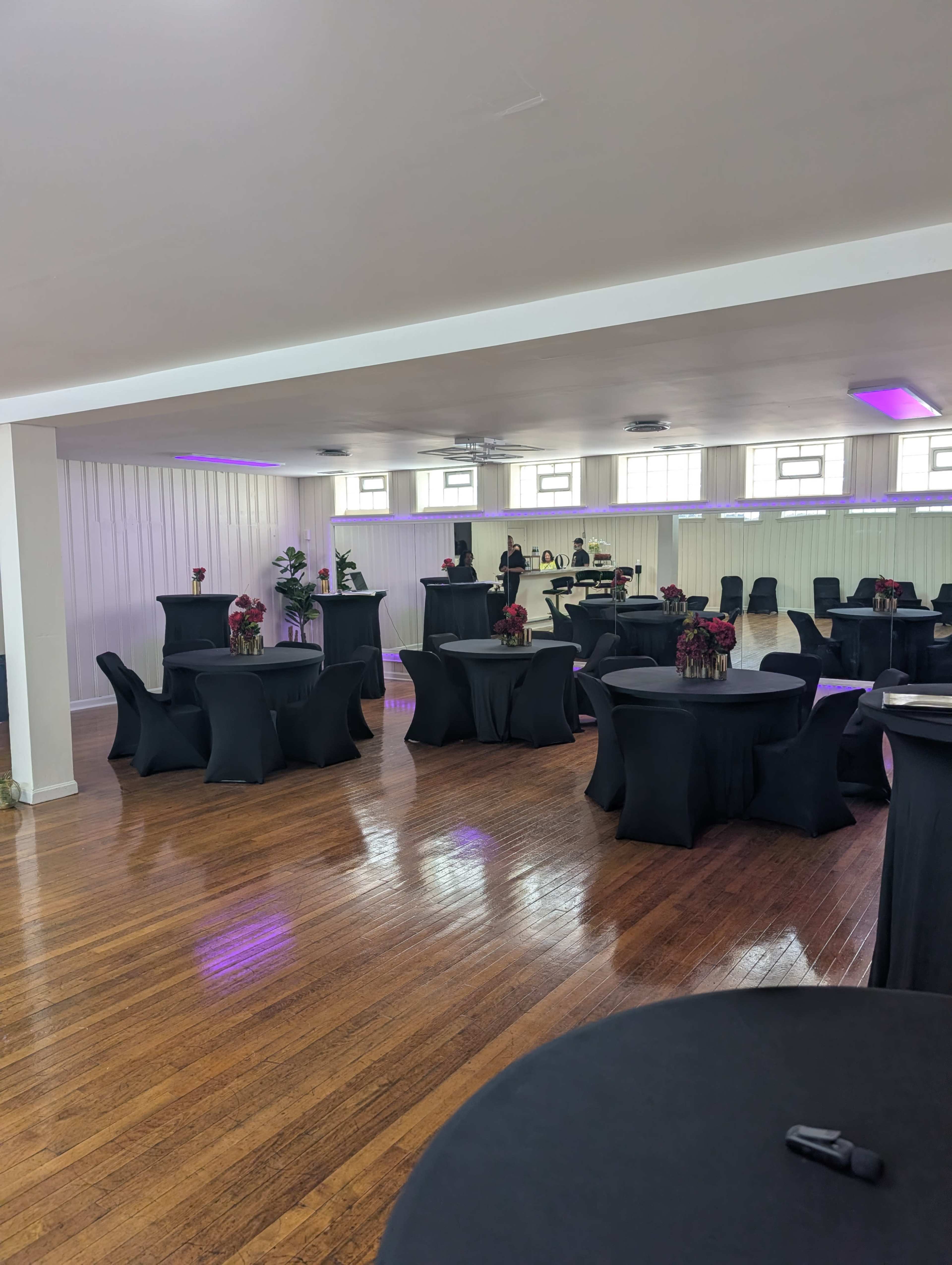 The image shows a spacious event venue featuring several round tables covered with black tablecloths, arranged neatly with floral centerpieces.