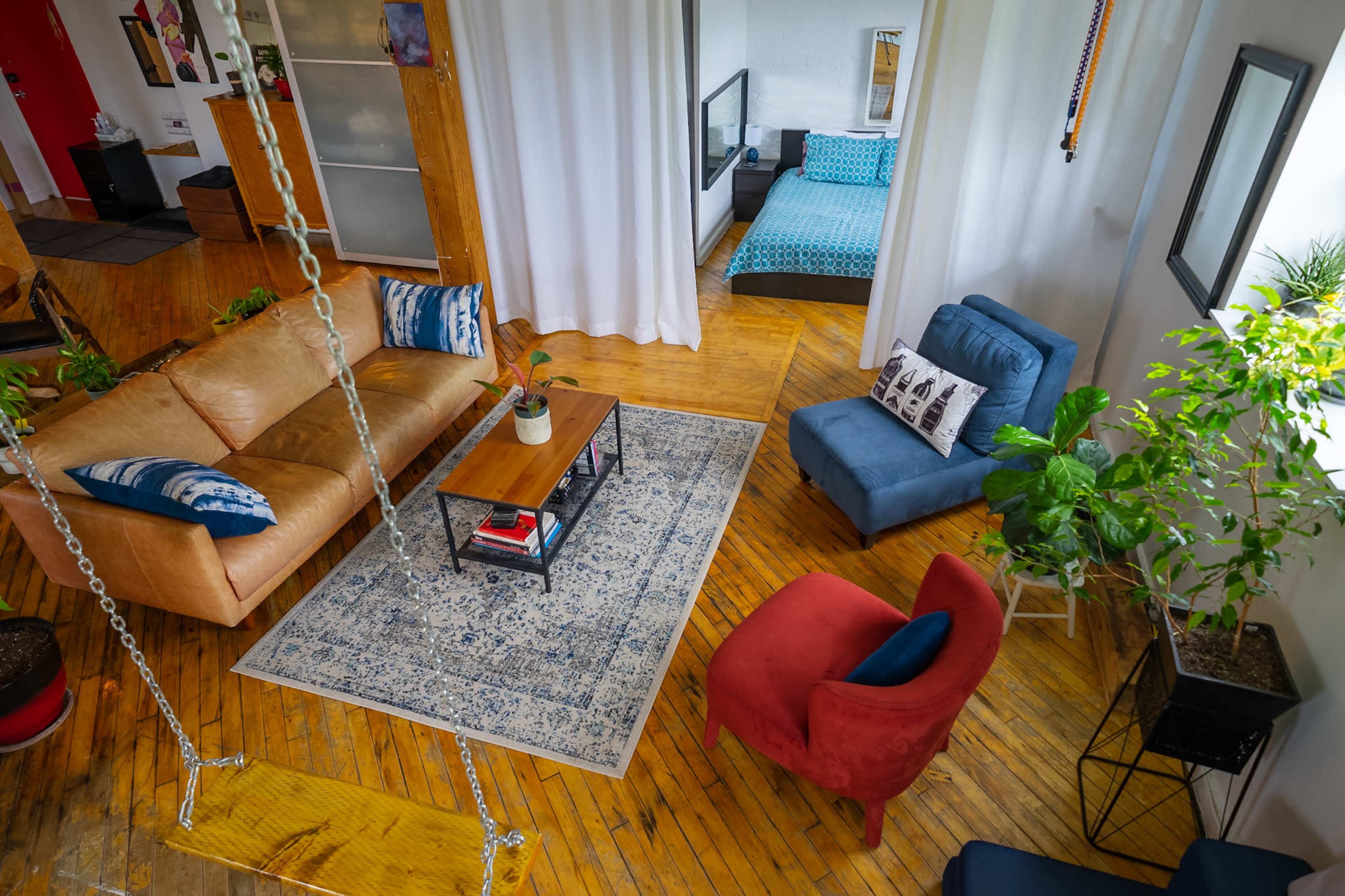The image shows a spacious living area with a brown sofa, a blue armchair, a red chair, a coffee table on a patterned rug, and plants, with a bedroom visible in the background.