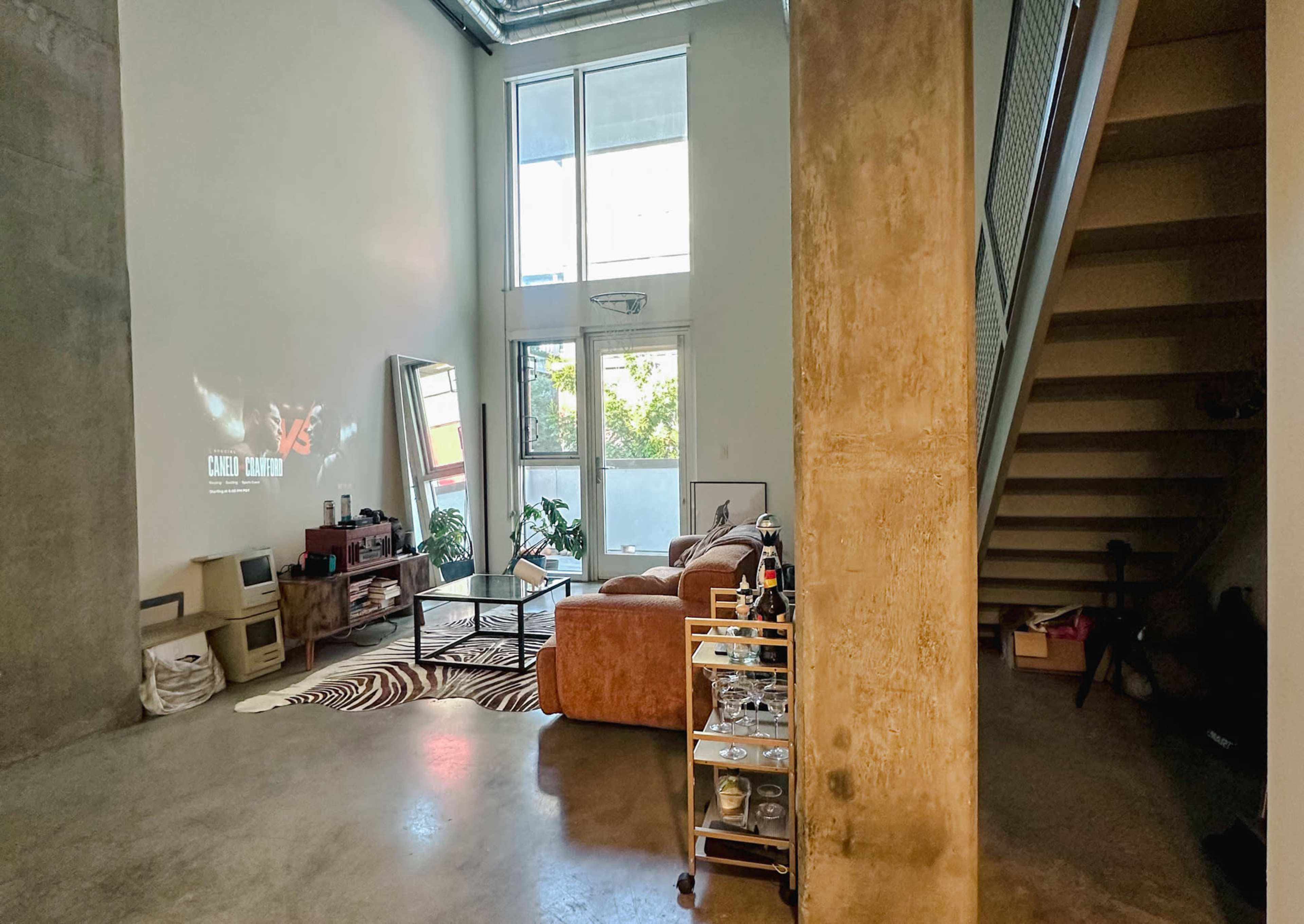 The image shows an industrial-style living space with a concrete wall, a cozy seating area, and a staircase leading to an upper level.