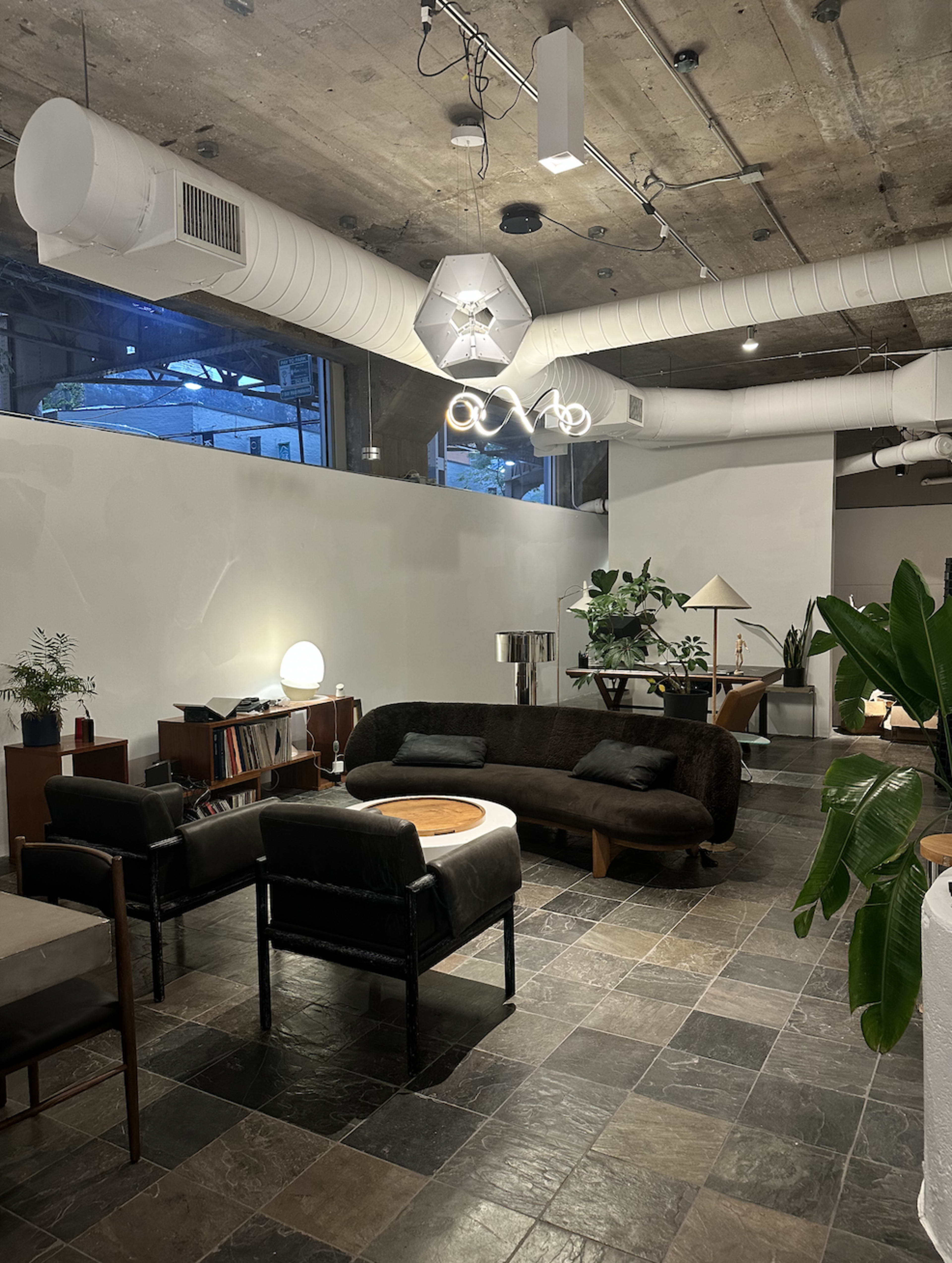 The image depicts a modern, minimalist interior space featuring a cozy seating arrangement with a dark sofa, armchairs, and various plants, illuminated by overhead lighting.