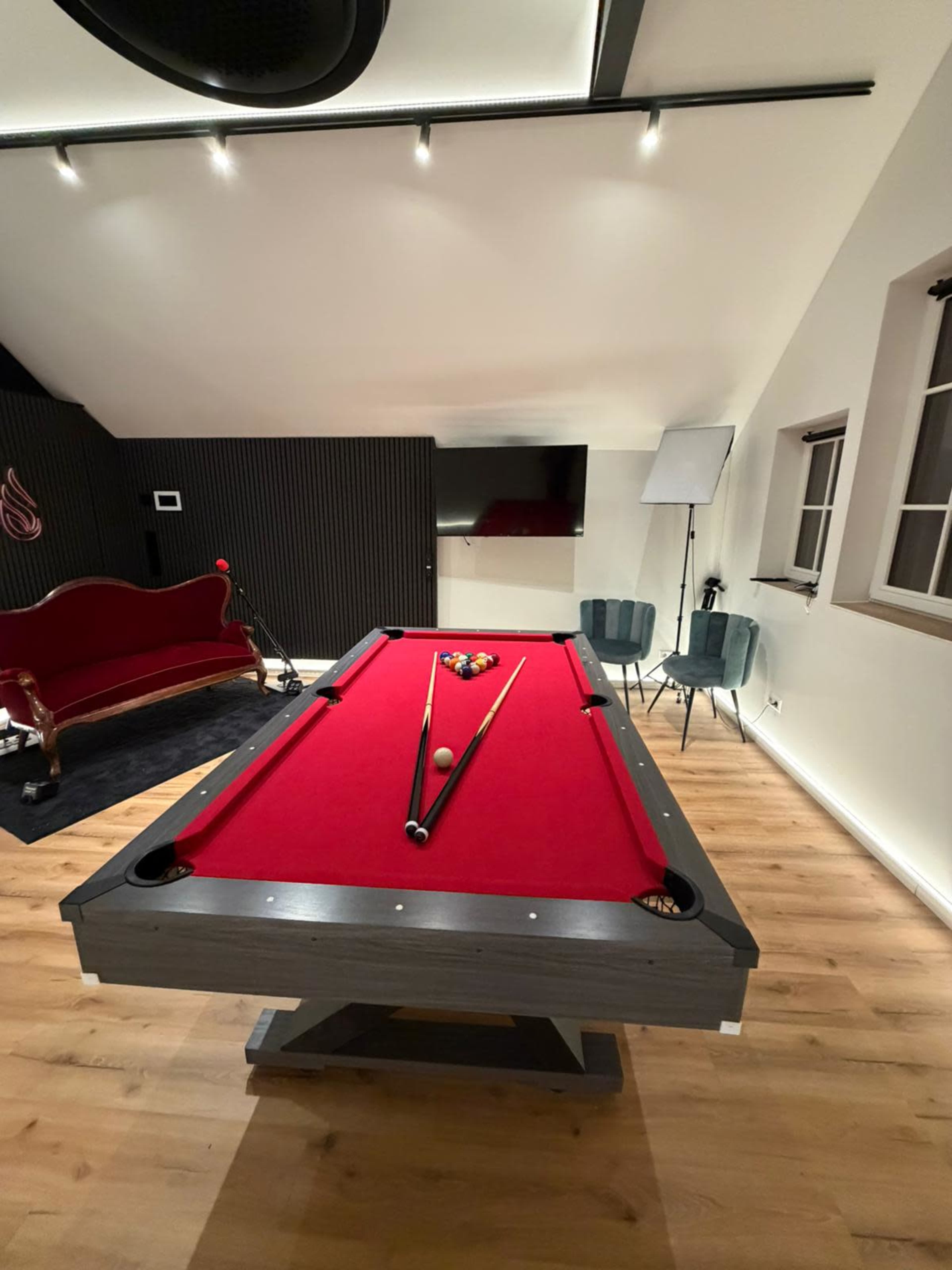The image shows a red felt pool table with cues and balls set in a sleek, modern room featuring wooden flooring and contemporary furniture.