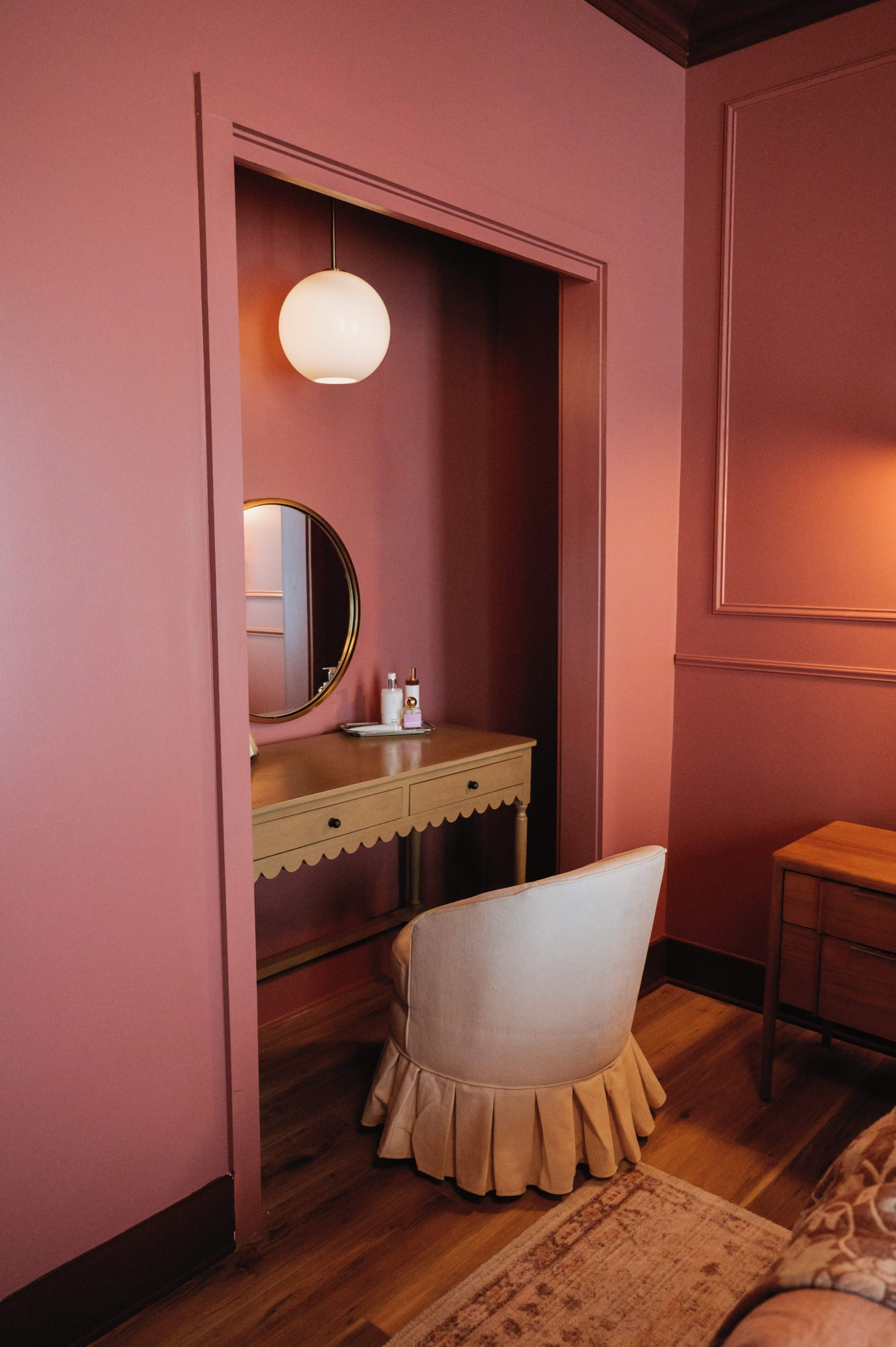 The image shows a small vanity area with a round mirror and a chair, all set against a pink-painted wall.