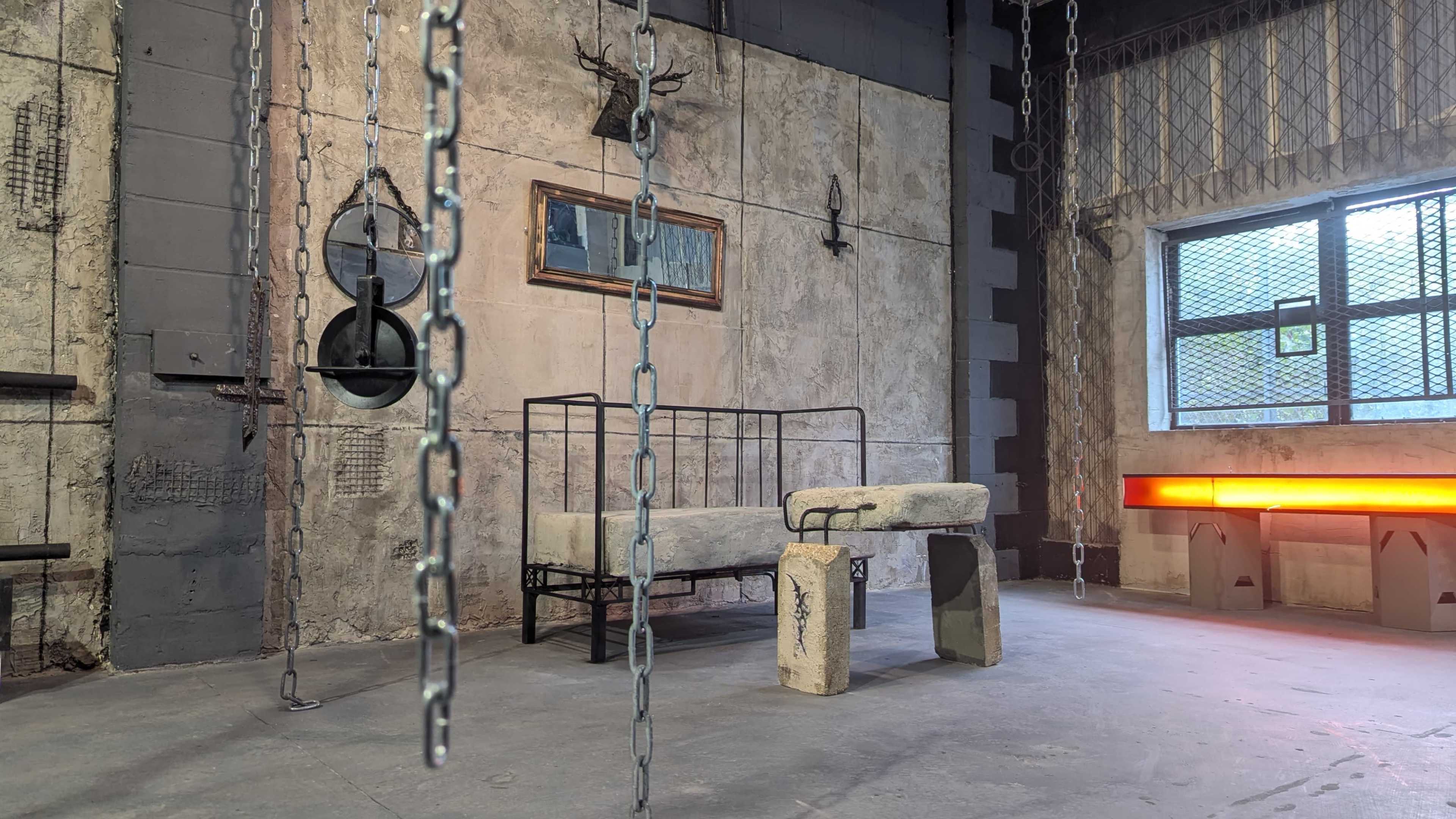 The scene features a stark, industrial room with a concrete floor, a bed frame, a stone bench, and hanging chains, set against a textured gray wall.