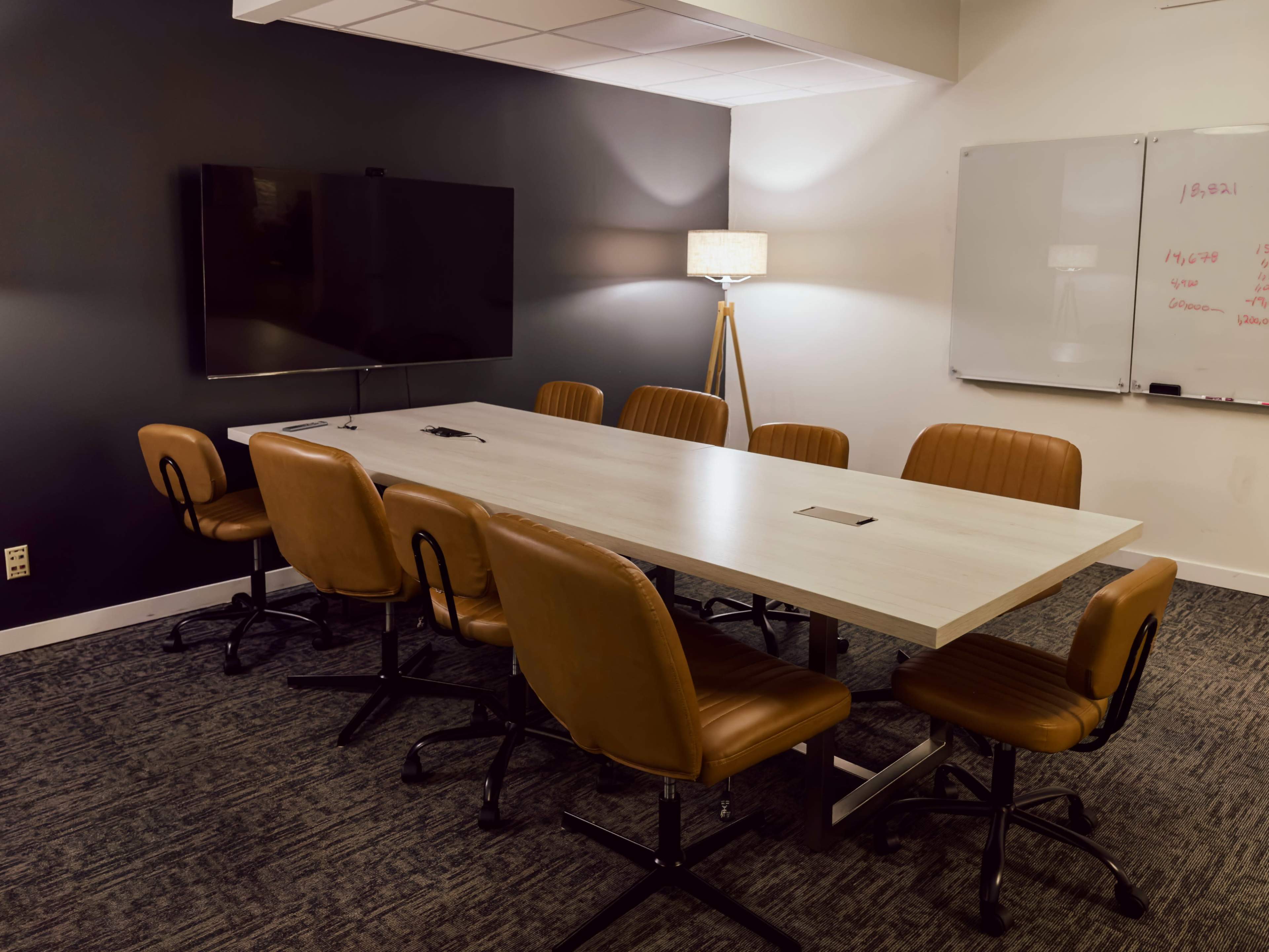 A conference room features a long white table surrounded by eight brown leather chairs, with a television display on one wall and a lamp in the corner.